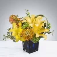 Cube Blue - This stunning floral arrangement, known as &quot;Cube Blue,&quot; is a delightful blend of vibrant yellow lilies and the unique pincushion protea. The architectural shape of the protea adds an intriguing dimension to the display, while the striking blue cube vase provides a modern and sophisticated touch. The combination of bright colors and sleek lines makes this arrangement an ideal gift for various occasions.