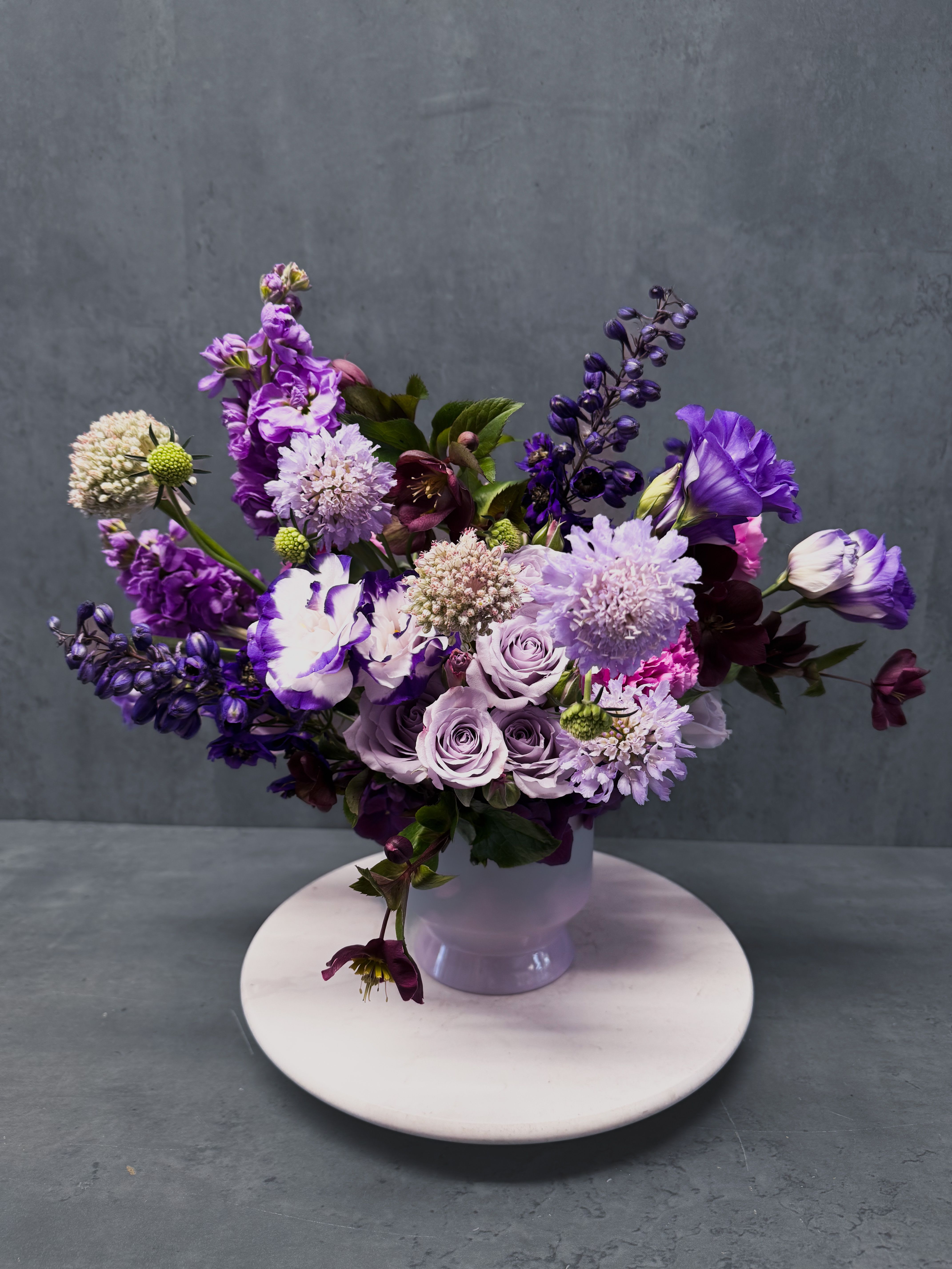 Lavender Haze Lavender Haze - A fun combination of purples and lavenders, in a ceramic compote vase. Made up of ornamental kale, stock, scabiosa and other seasonal florals. **All arrangements will vary slightly based off seasonal availability. **