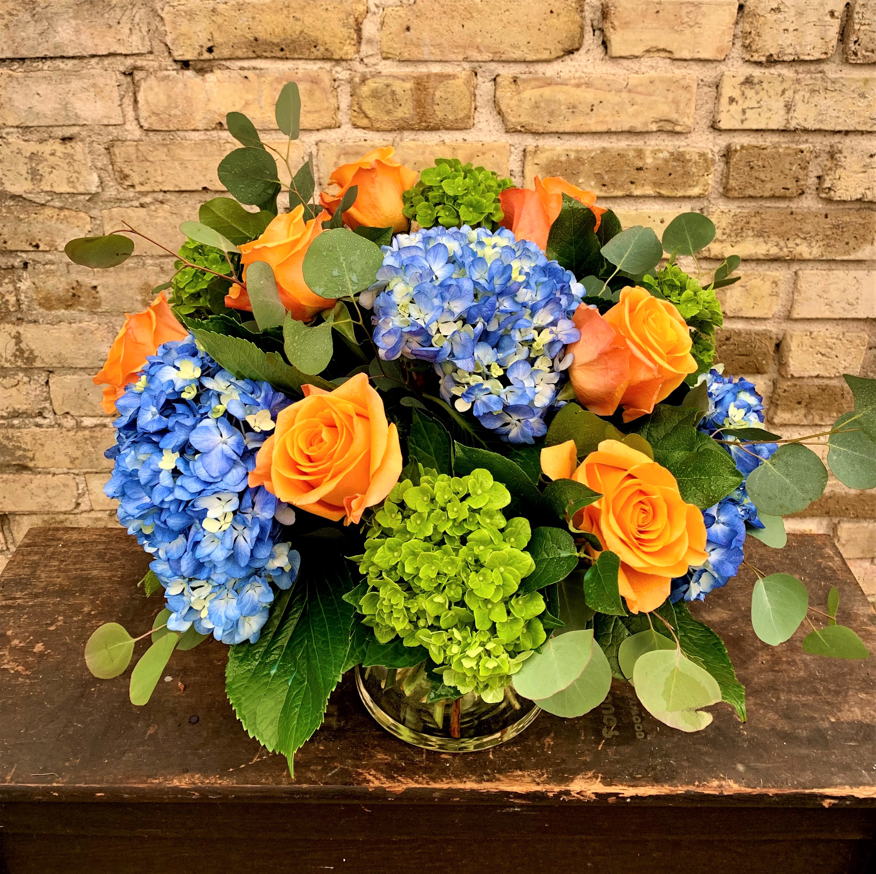 East Coast Design - Fresh Arrangement Designed in a Clear Vase Using Blue and Green Hydrangea, Orange Roses and  Mixed Foliage's. Color of Rose May Vary.