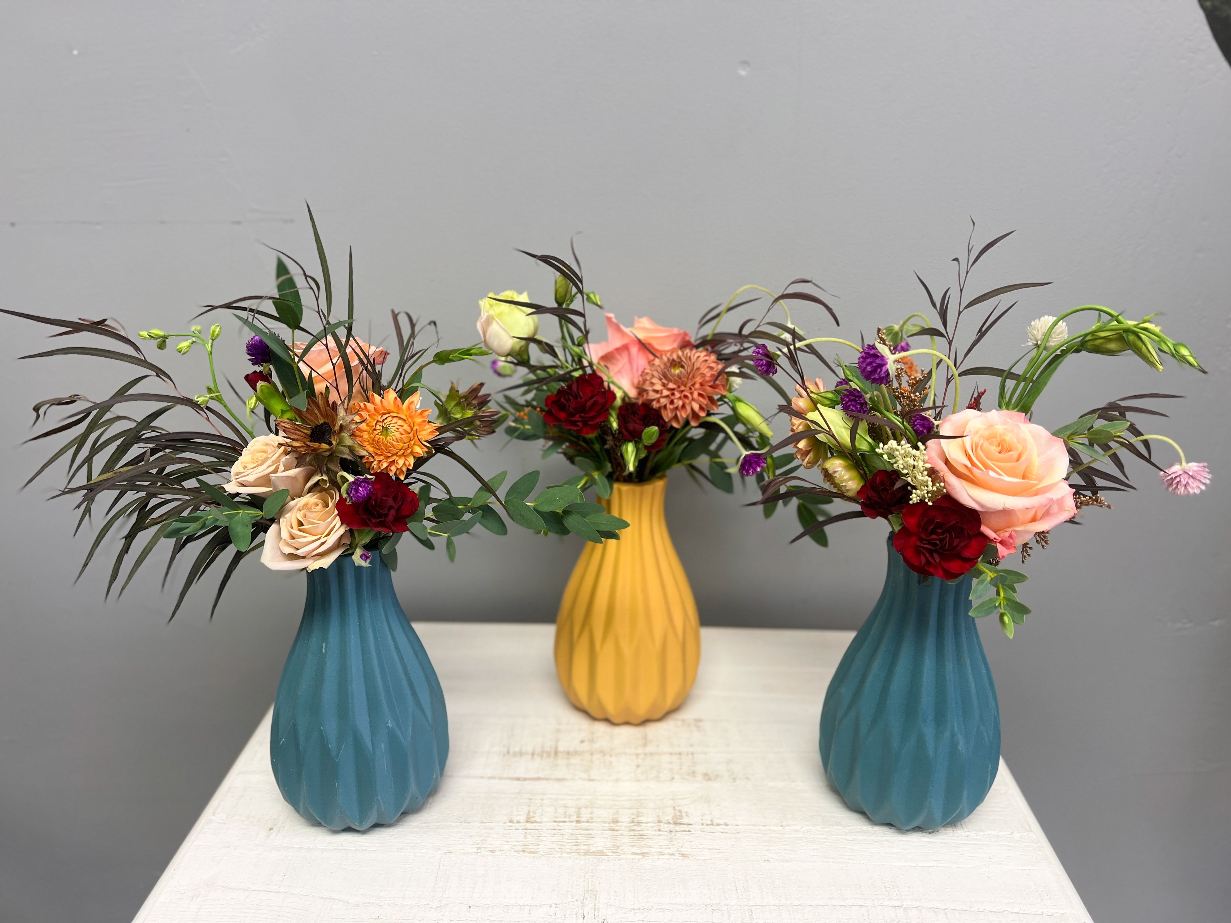Autumn Radiance Vase Trio Autumn Radiance Vase Trio - A vibrant mix of roses, mums, and textural blooms styled in sculpted teal and mustard ceramic vases. Choose one or a trio — each one artfully arranged and uniquely yours. autumn floral arrangement, modern vase flowers, teal vase bouquet, mustard vase arrangement, fall flower décor, small table flowers, colorful floral trio, handcrafted flower arrangement, unique vase design, Jackson TN florist, Anointed Flowers gifts, local flower delivery