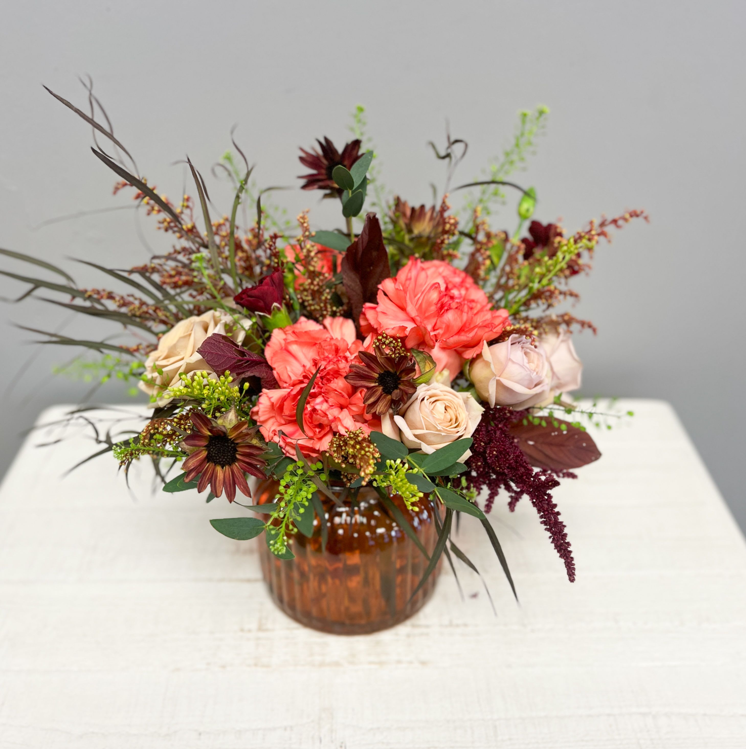 Amber Blooms - Warm, vibrant, and full of fall charm — the Amber Glow arrangement blends the richness of autumn tones with soft romantic blooms. Designed in a ribbed amber glass vase, this seasonal favorite features a lush mix of coral carnations, mauve and tan roses, rust daisies, amaranthus, and textured greens that create a natural, gathered look.  Perfect as a centerpiece for your table, a thoughtful gift, or an elegant accent for any space. Each arrangement is hand-designed with premium seasonal flowers, making every piece uniquely yours.