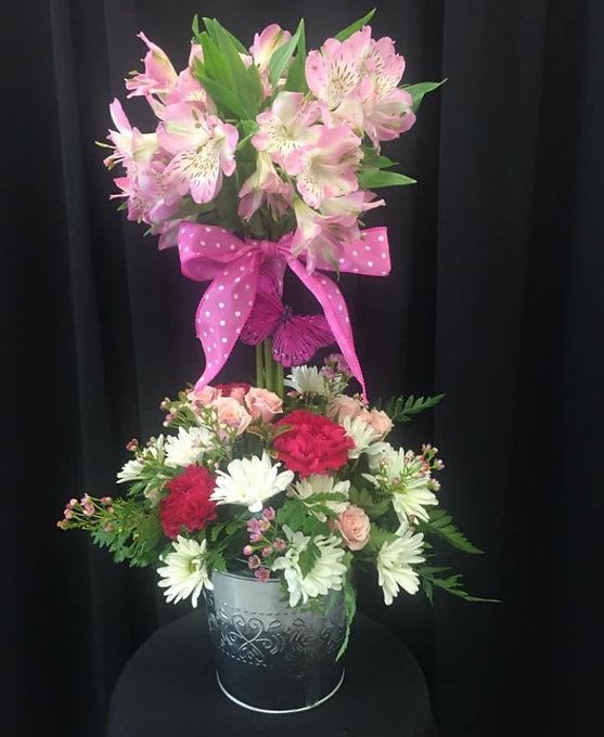 Topiary Tree Topiary Tree - "Sweet Surprise" Arrangement This cheerful two-tiered floral display is bursting with personality! Featuring soft pink alstroemeria blooms standing tall above a garden-fresh mix of white daisies, rich red carnations, blush spray roses, and delicate filler flowers, all nestled in an embossed metal container. Finished with a playful pink polka dot bow and butterfly accent, this arrangement is perfect for birthdays, celebrations, or just because.