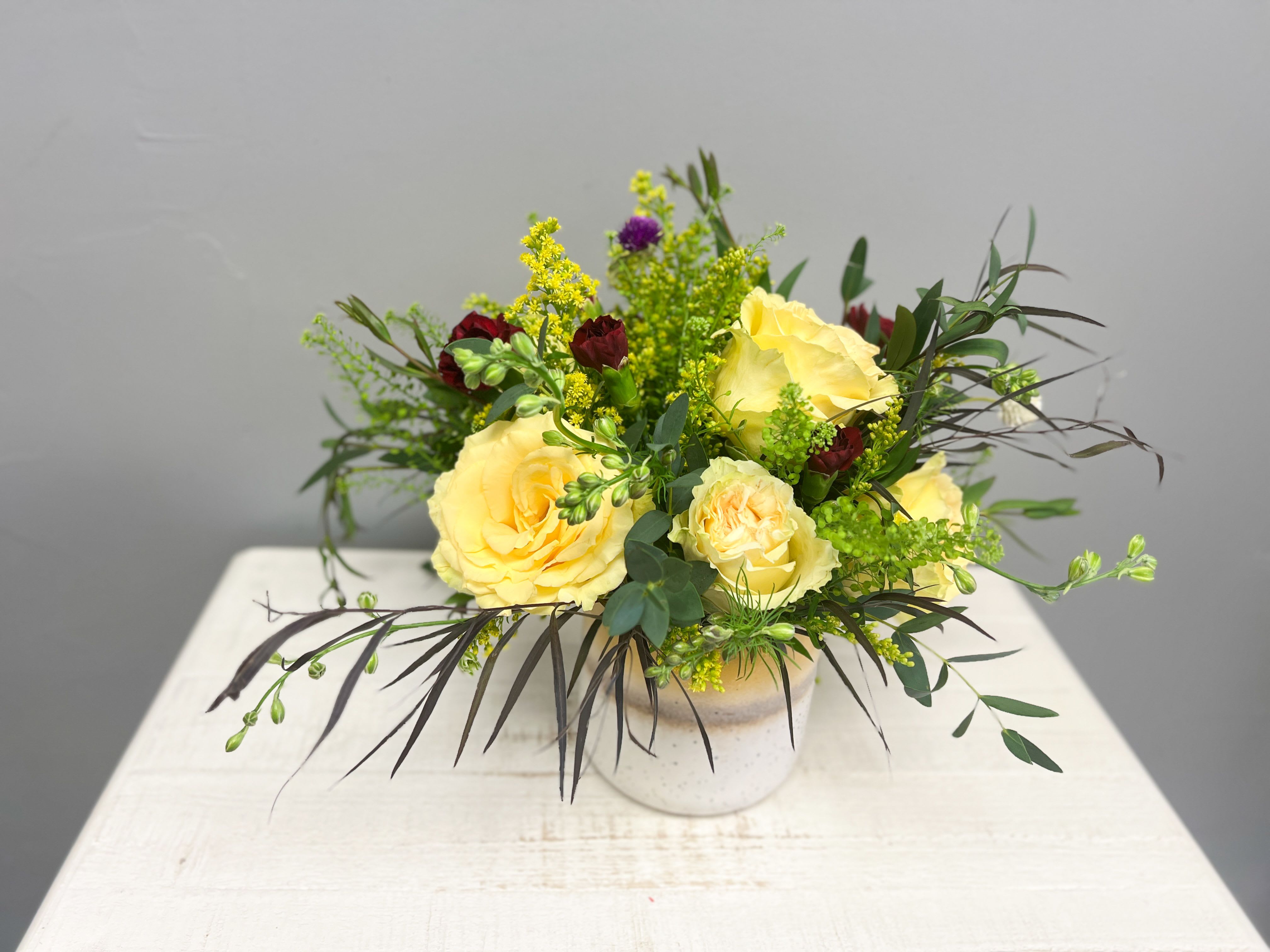Golden Hour - Radiating warmth and happiness, the Golden Hour arrangement brings sunshine to any space. Designed in a modern speckled ceramic vase, this fresh mix of blooms features buttery yellow roses, deep burgundy accents, and pops of greenery that create a perfect harmony of light and contrast.  Ideal for brightening a home, celebrating a special occasion, or sending a cheerful “thinking of you” surprise — this arrangement captures the glow of golden hour in floral form.  