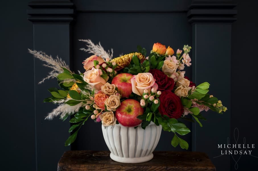 Apple Crisp - A warm and inviting arrangement featuring Sahara spray roses, bronze Cremones, cream Hypericum berries, jasmine vine greenery, pampas grass, peach shimmer roses, orange ranunculus, red heart roses, peach stock, and fresh apples, artfully combined to create a rich, seasonal display.