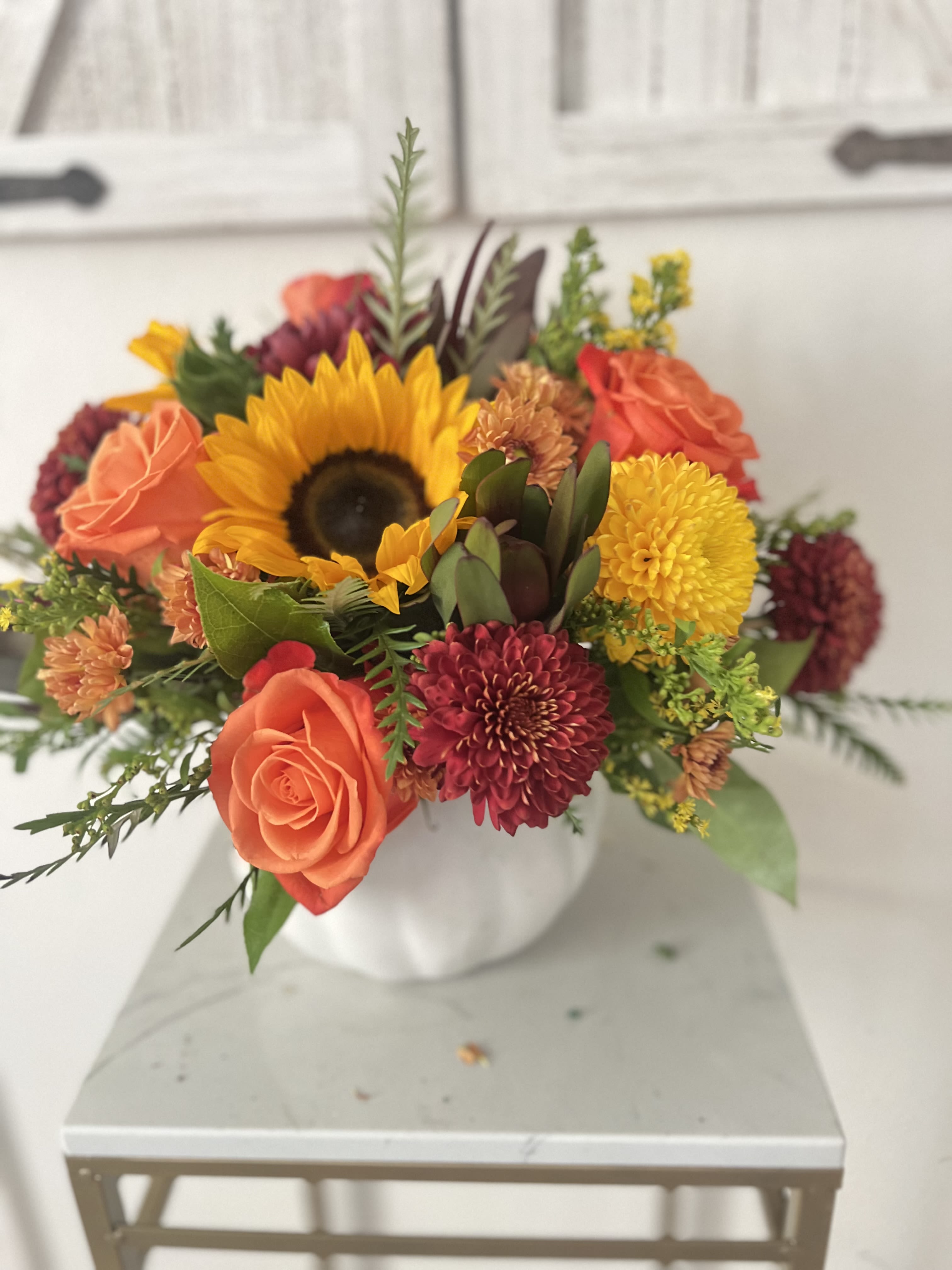  Pumpkin Harvest Glow -  A radiant mix of autumn blooms bursts from a charming white pumpkin vase. Featuring bright sunflowers, vivid orange roses, and deep burgundy mums, Pumpkin Harvest Glow celebrates the warmth and richness of the fall season. It’s the perfect piece to brighten your table or bring cozy harvest vibes into your home.