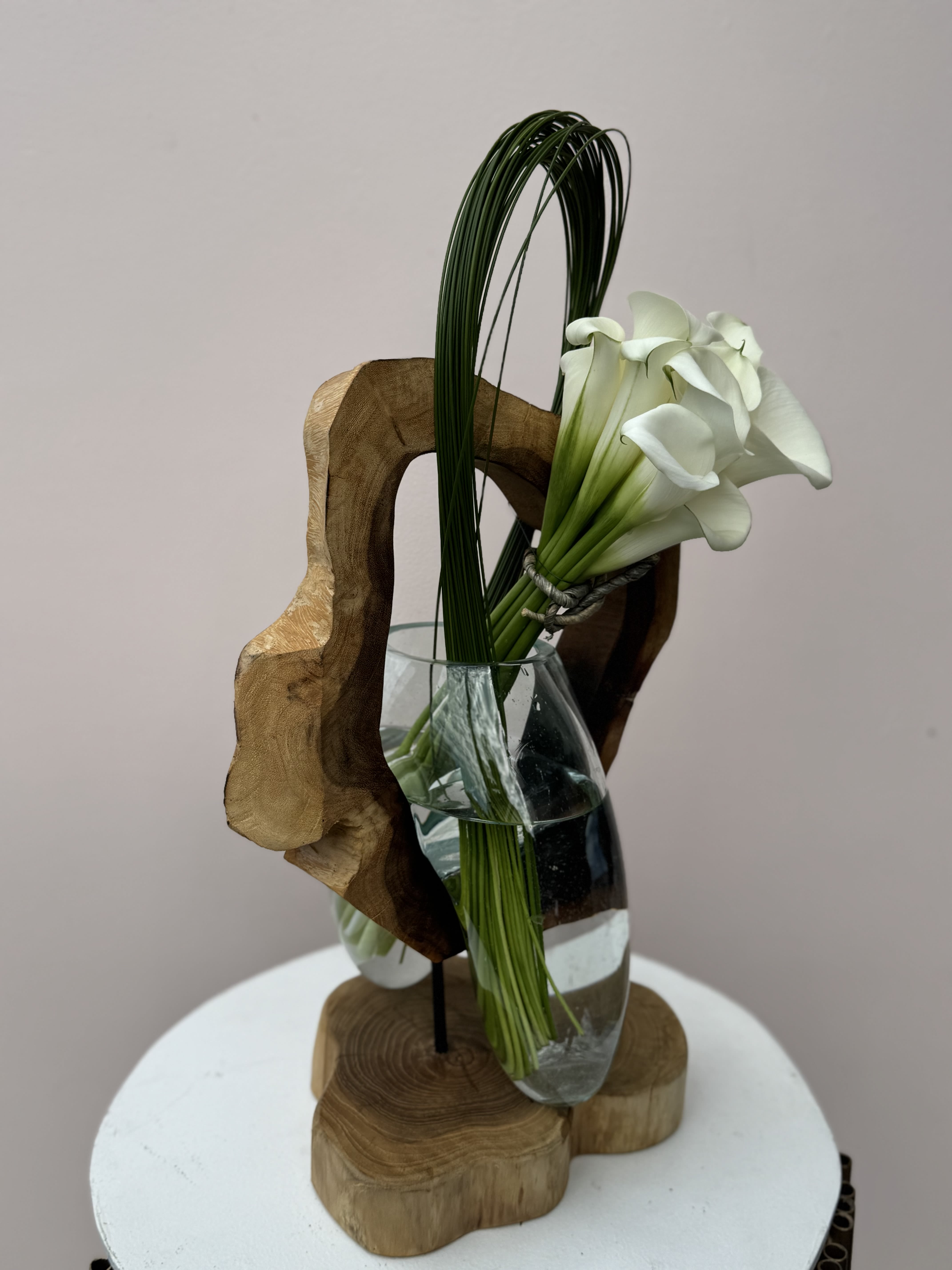 Echos of Lily - A sleek floral arrangement featuring elegant white calla lilies artfully displayed in a clear glass vase, anchored within a minimalist wooden structure. This design blends natural textures with refined simplicity—perfect for contemporary interiors.