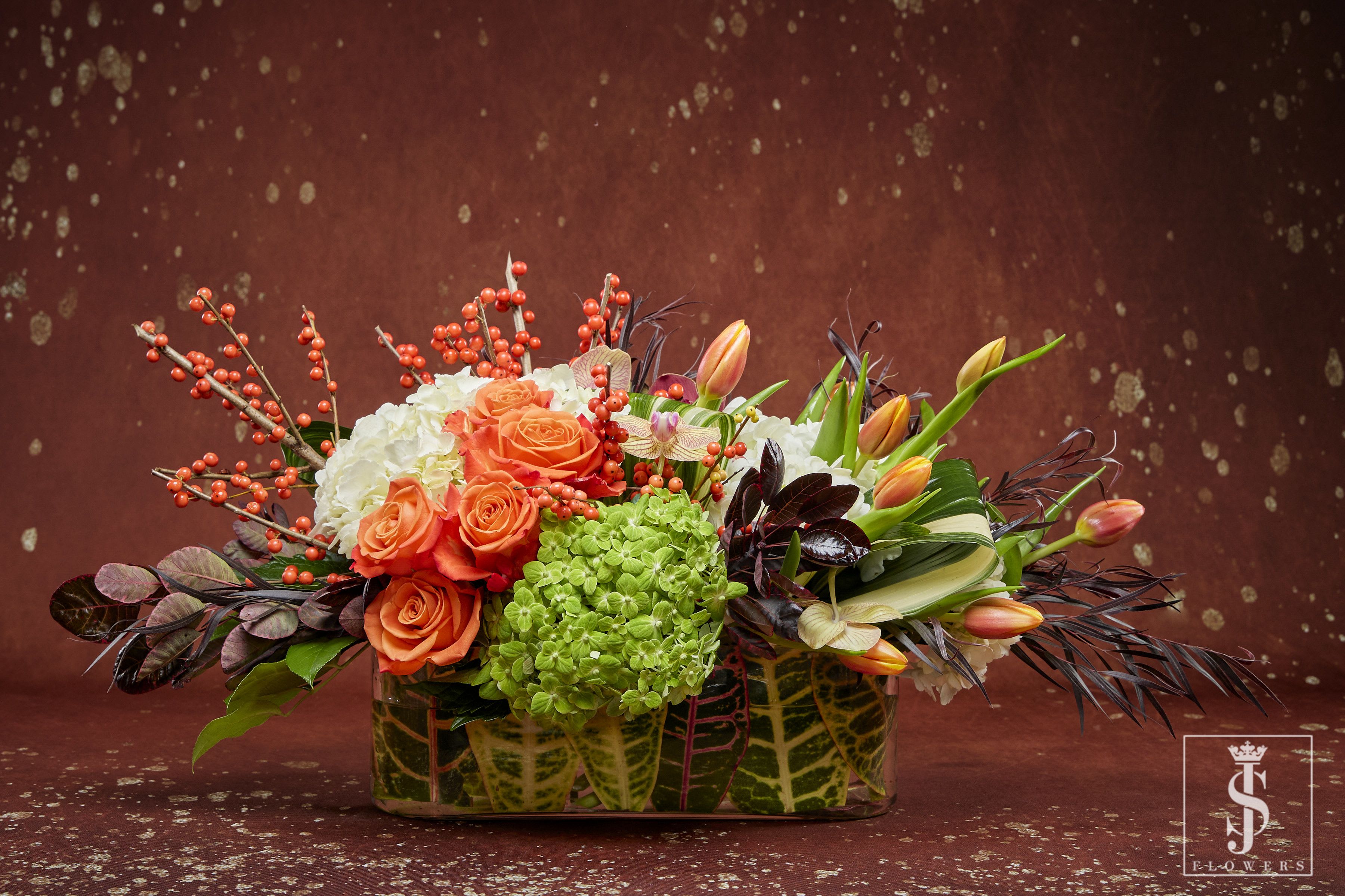 Amber Glow Long and Low Centerpiece - Capture the warm glow of autumn with this rich, amber-toned arrangement. Featuring Orange roses, white &amp; green hydrangeas, Tulips, orchids, it’s accented with asclepia and mini Italian pitt in a clear glass vase. 