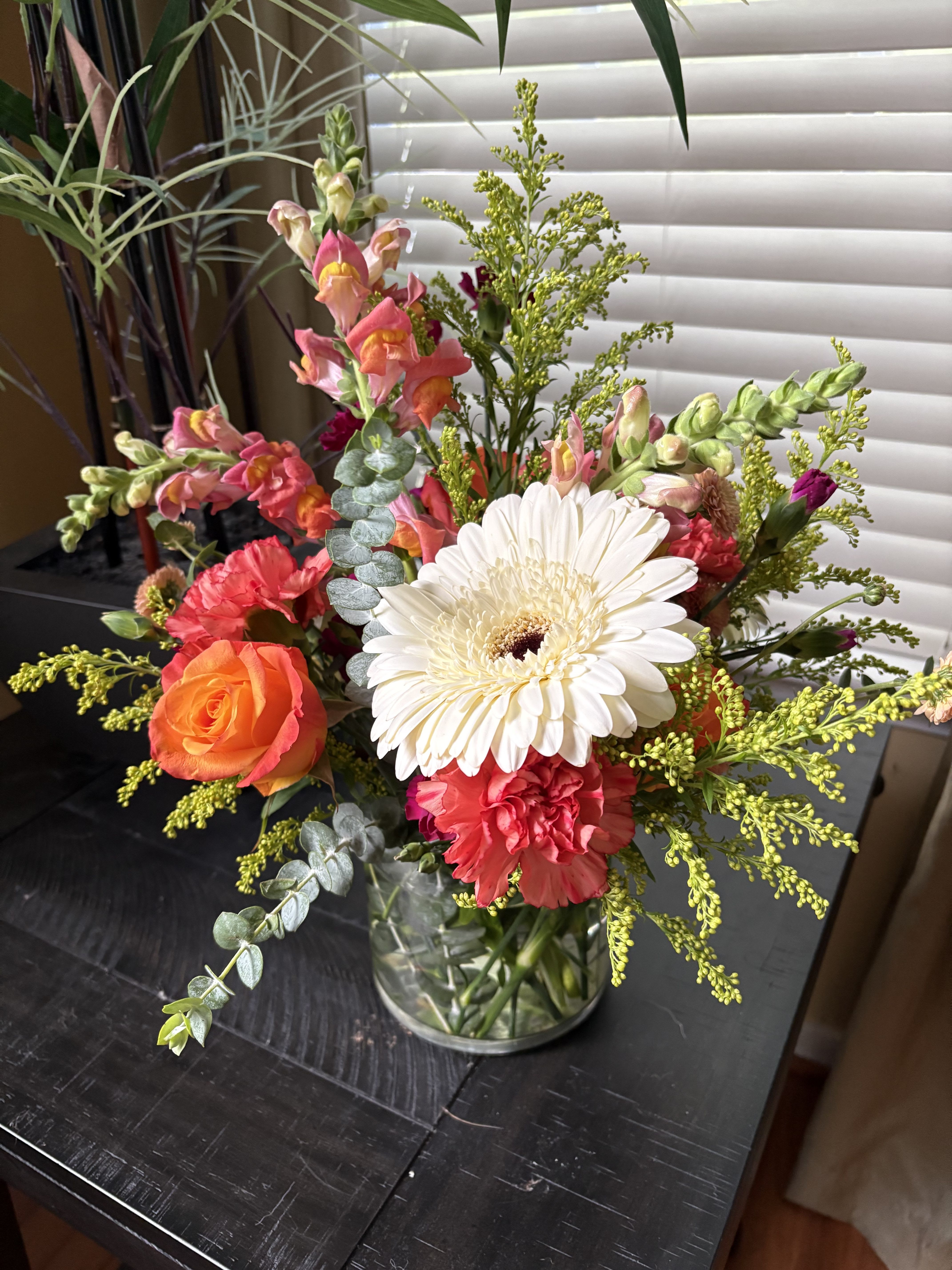 Autumn Radiance - Celebrate the vibrant beauty of the season with Autumn Radiance — a stunning mix of warm and cheerful blooms arranged to brighten any space. This lively design features white gerbera daisies, coral roses, red carnations, and playful pink snapdragons, accented with golden solidago and lush eucalyptus for texture and balance. Displayed in a clear glass vase, this arrangement perfectly captures the cozy charm and colorful spirit of fall.   Ideal for birthdays, thank-you gifts, or simply adding a burst of joy to your home decor.