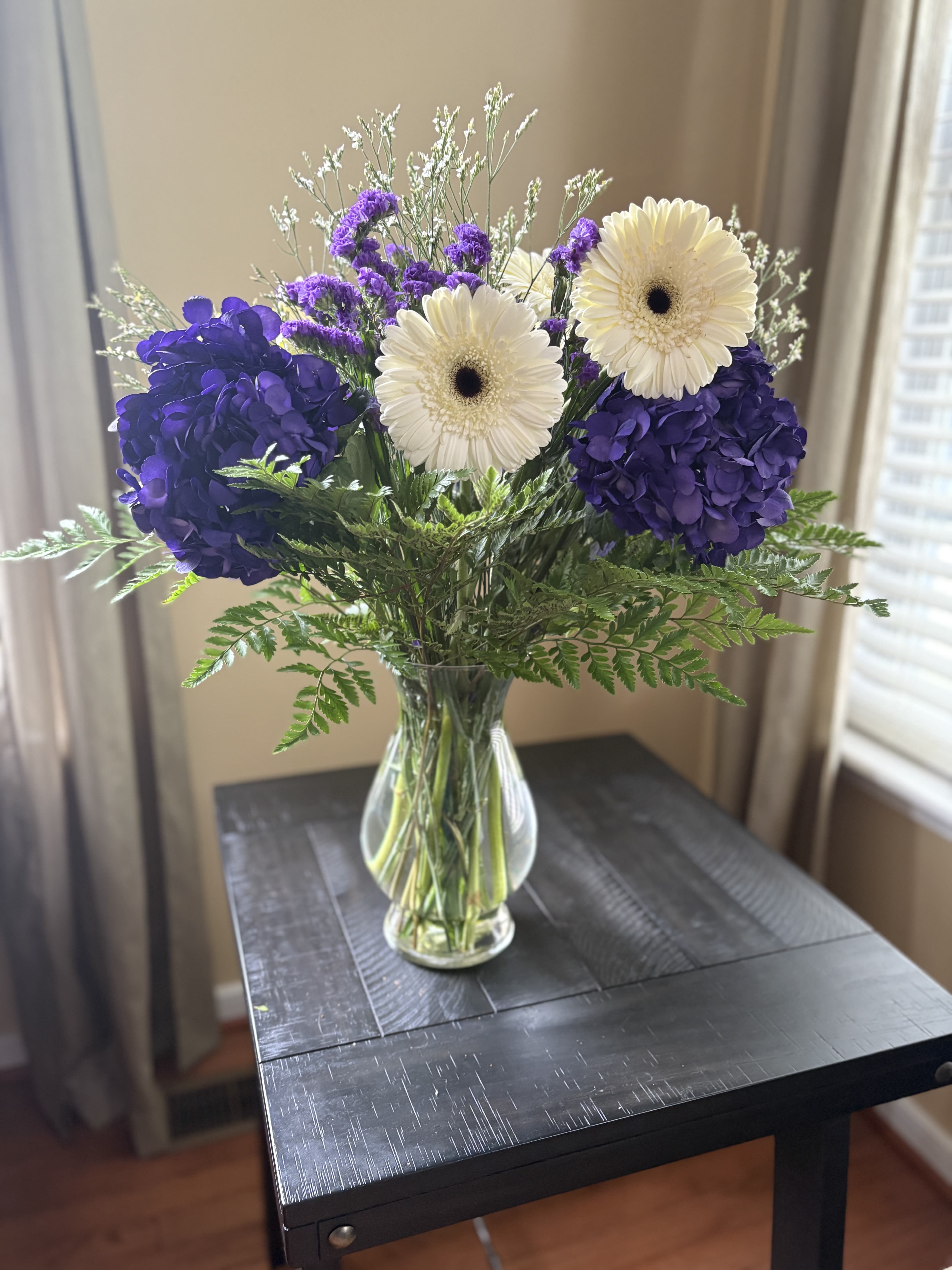 Lavender Breeze - Fresh. Uplifting. Gracefully Chic.  Let elegance bloom with “Lavender Breeze”, a stunning arrangement featuring lush purple hydrangeas paired with crisp white Gerbera daisies, delicately arranged in a clear glass vase. This beautiful blend of soft textures and gentle hues creates a soothing, yet joyful display — perfect for birthdays, get-well wishes, thank you gifts, or simply brightening someone’s day.  With its calming color palette and timeless design, “Lavender Breeze” brings a breath of fresh air to any space.  Includes: Premium Purple Hydrangeas Fresh White Gerbera Daisies Accented with Seasonal Greenery Arranged in a Glass Vase