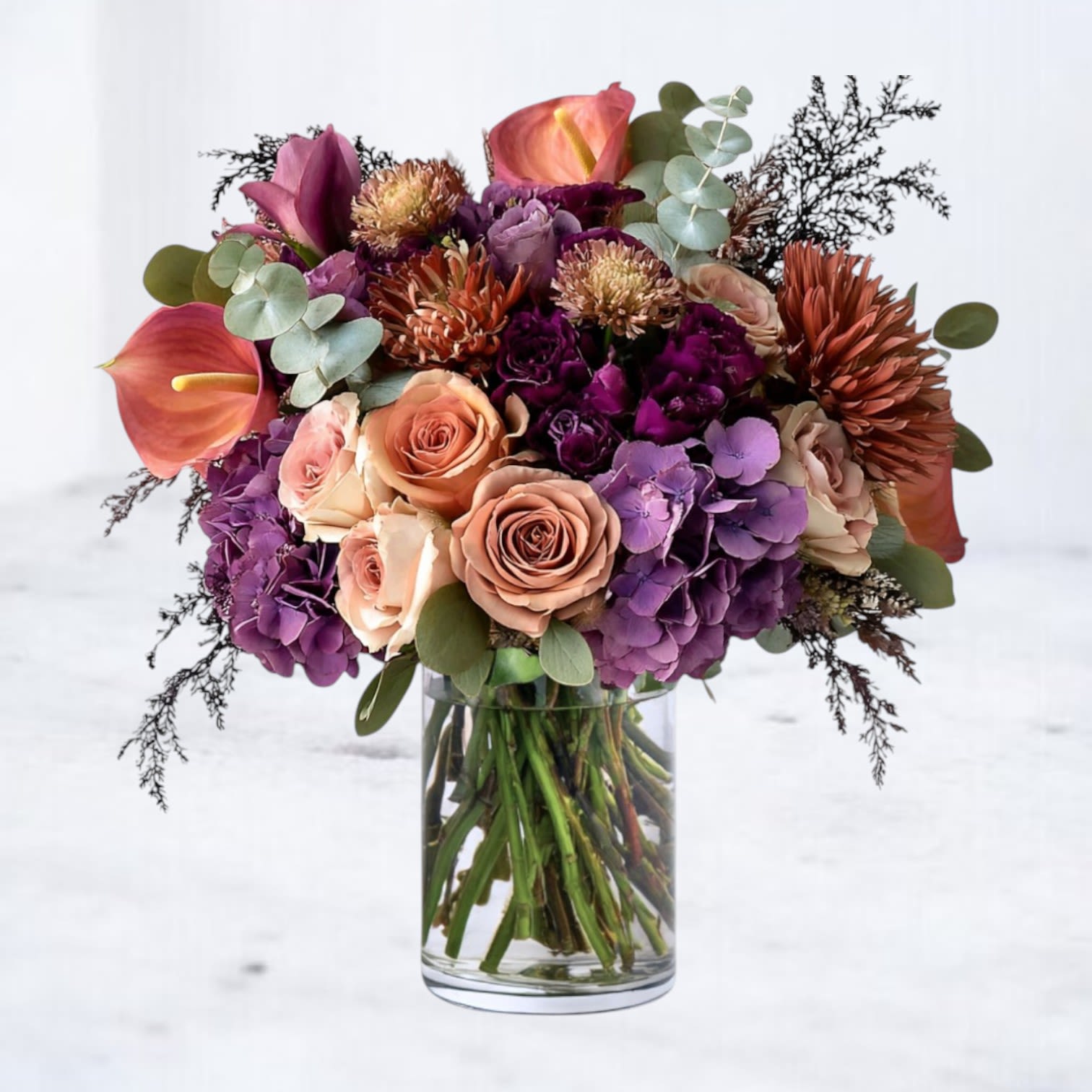 Duskmere Duskmere - Step into the twilight beauty of the Duskmere Bouquet, a captivating arrangement that captures the quiet allure of fall evenings. Warm peach roses, deep plum hydrangea, copper mums, and burnt-orange calla lilies intertwine with textured greenery and soft mauve blooms, creating a palette that feels both modern and timeless. Designed in a sleek glass vase, this luxurious composition exudes depth, elegance, and a hint of mystery — perfect for birthdays, romantic gestures, or sophisticated autumn gatherings.