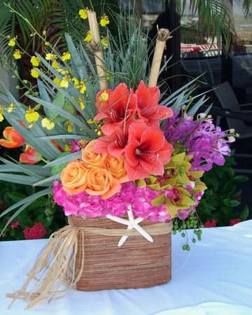 Splash of Paradise -  Size: Standard: A small arrangement measuring approximately 12-15″ in width. Grouping of orange roses, pink and yellow orchids with big blooms of coral amaryllis, hot pink hydrangeas ,tulips and tropical foliage . Accent of bamboo, starfish and raffia as a natural elements . 