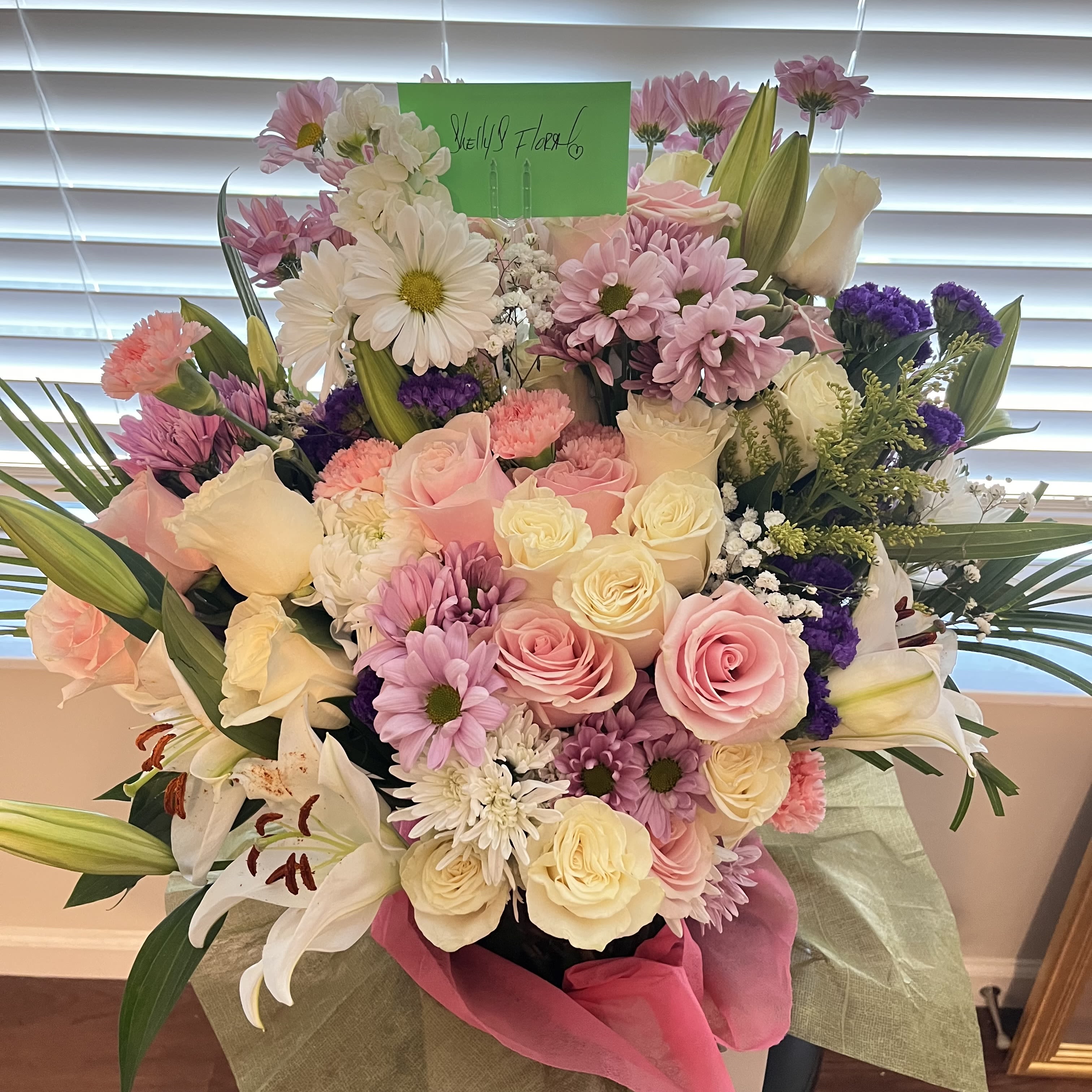 Blushing Elegance By Shelly - This radiant design features soft pink and ivory roses, cheerful daisies, delicate carnations, vibrant mums, and fragrant lilies, accented with lush greenery and fillers. Overflowing with texture and color, this arrangement celebrates beauty, love, and joy—perfect for any occasion where words alone aren’t enough.   Handcrafted with care by Shelly’s Floral, every detail is arranged with intention to ensure freshness and elegance from vase to delivery.  Because every occasion deserves something this beautiful.