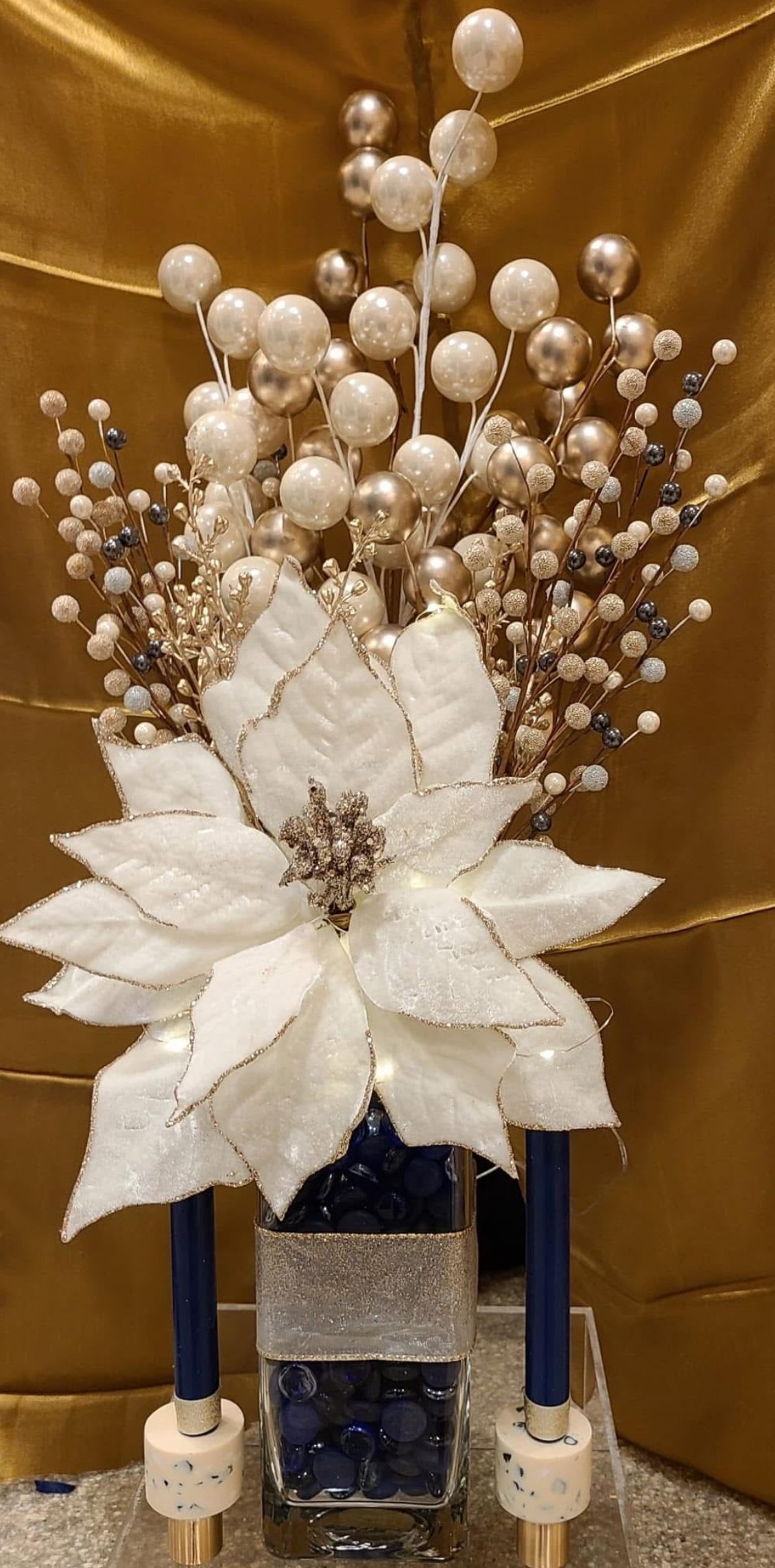 Dazzling Beauty Blue by Shelly's Floral - An exquisite centerpiece that captures timeless luxury and festive charm. Featuring shimmering pearl accents, golden details, and a bold white poinsettia, this handcrafted arrangement is designed in a modern glass vase filled with navy stones and finished with a soft metallic ribbon wrap. Flanked by coordinating taper candles, this piece makes a sophisticated statement for the holidays or as an all-season display of elegance.  Perfect for dining tables, mantels, or entryways, this arrangement is a lasting treasure that brings beauty and brilliance to any space. *Candles not included