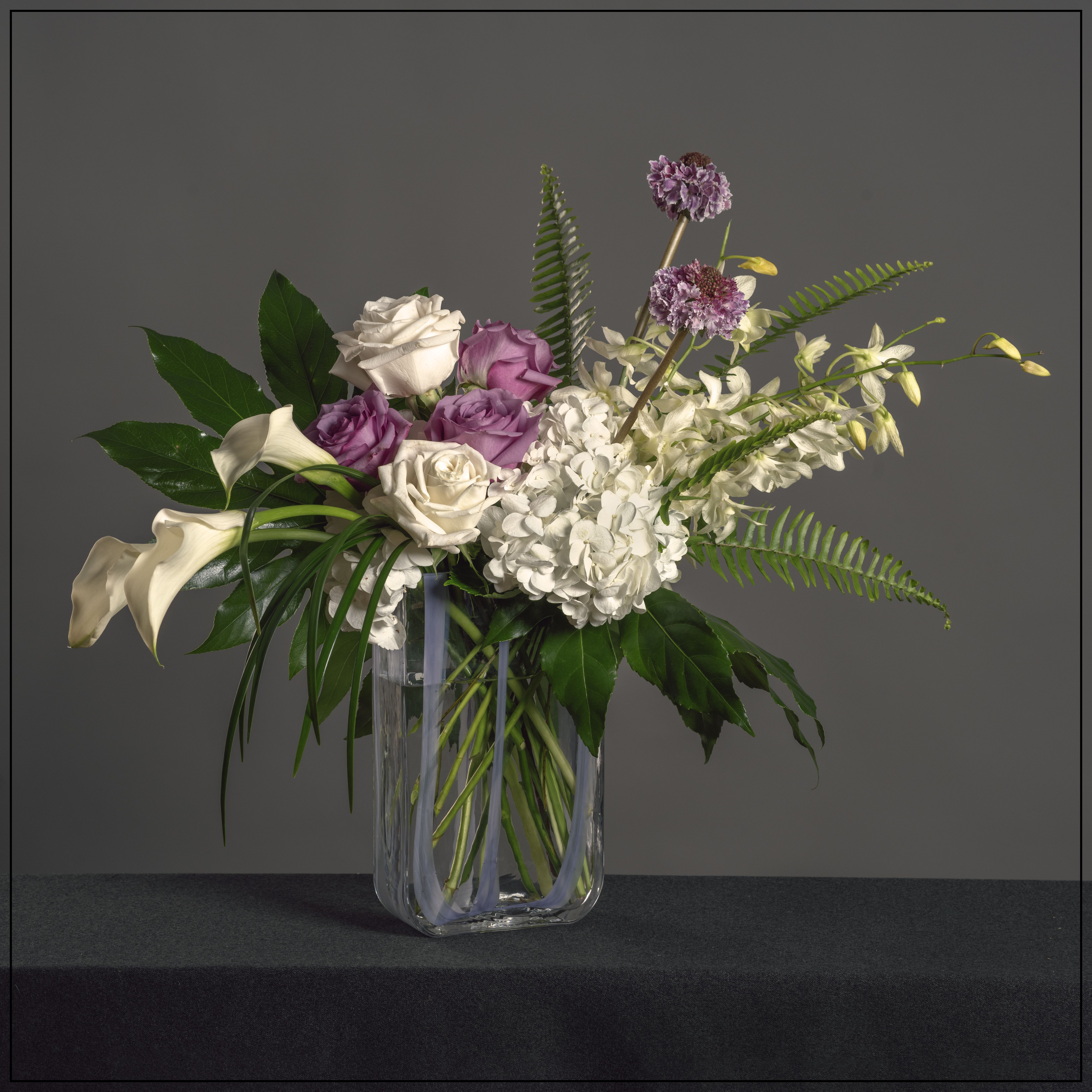 Katness Katness - An elegant mix of hydrangea. calla lily, roses, orchids and scabiosa with mixed tropical greens in a tall oblong vase. Dimensions - 20" H x 22" W
