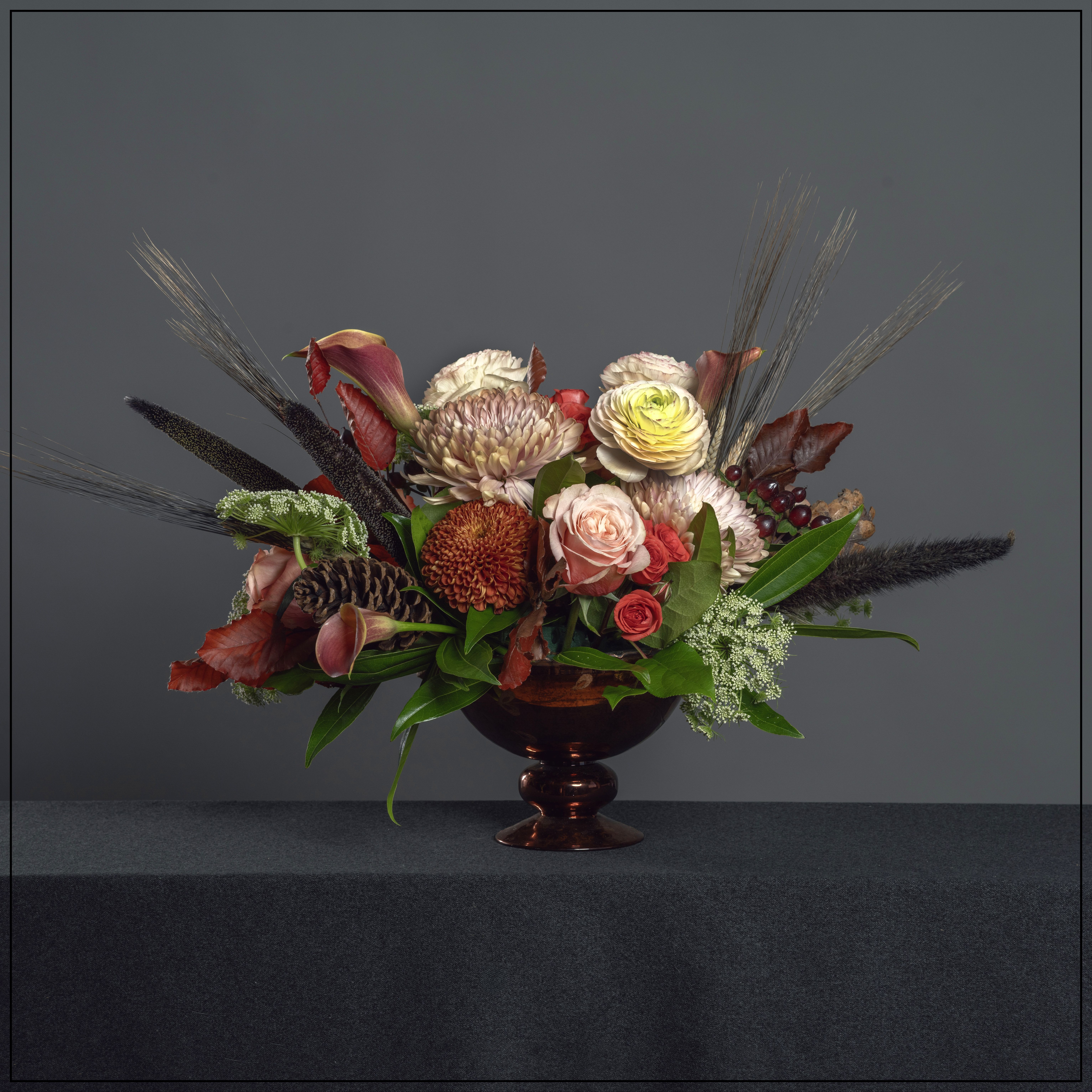 The Holiday Table The Holiday Table - Autumn is in the air with copper colored glass footed bowl full of ranunculus, calla lily, roses, hypericum berries, cremons with dried elements like pine cones, wheat and fall leaves. Dimensions: 14" H x 23" W