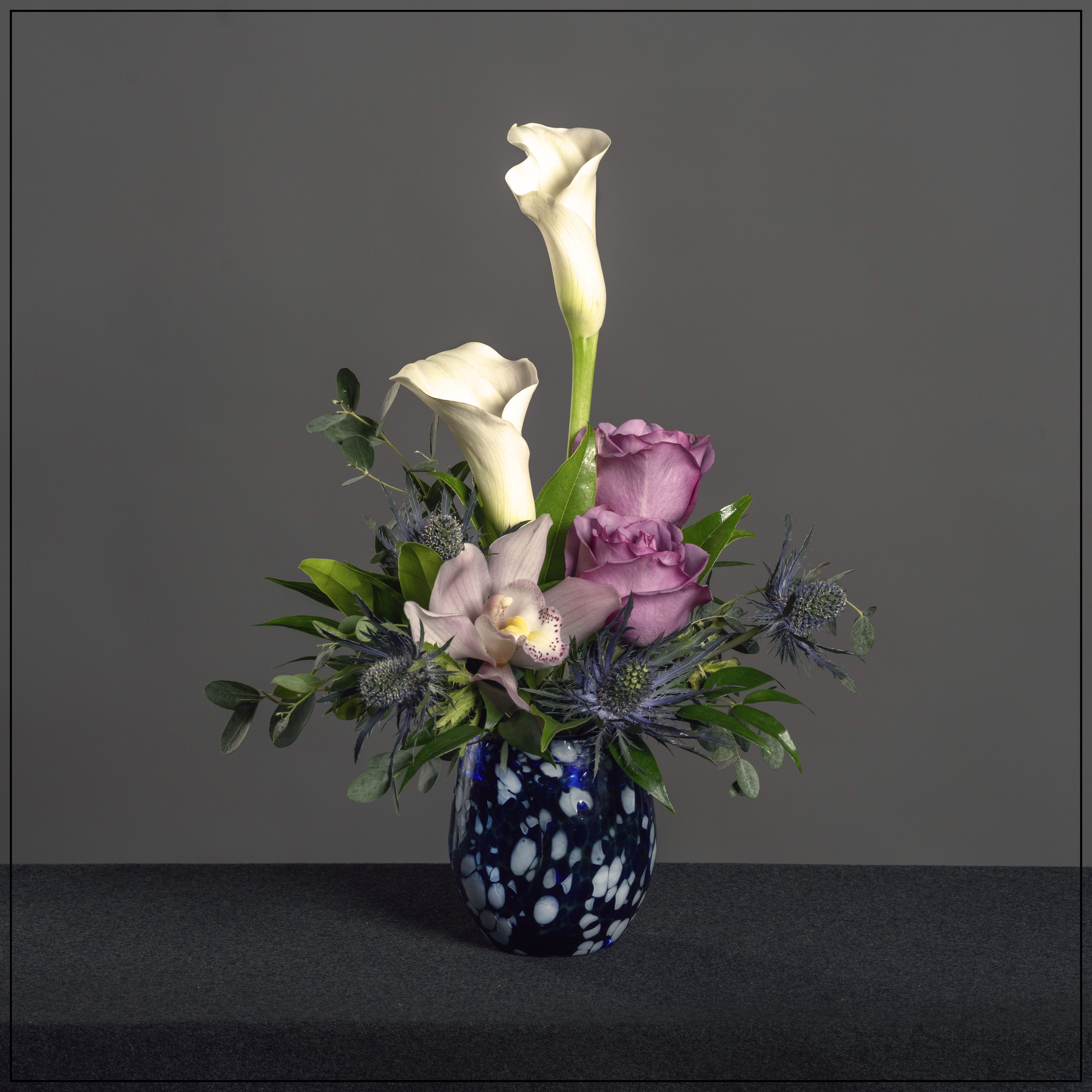 Blue Madeline Blue Madeline - This seasons version of our friend Madeline...In a art glass blue vase (art glass vase are all different) with calla lily, roses, orchid and eryngium. Dimensions: 15" H x 11" W