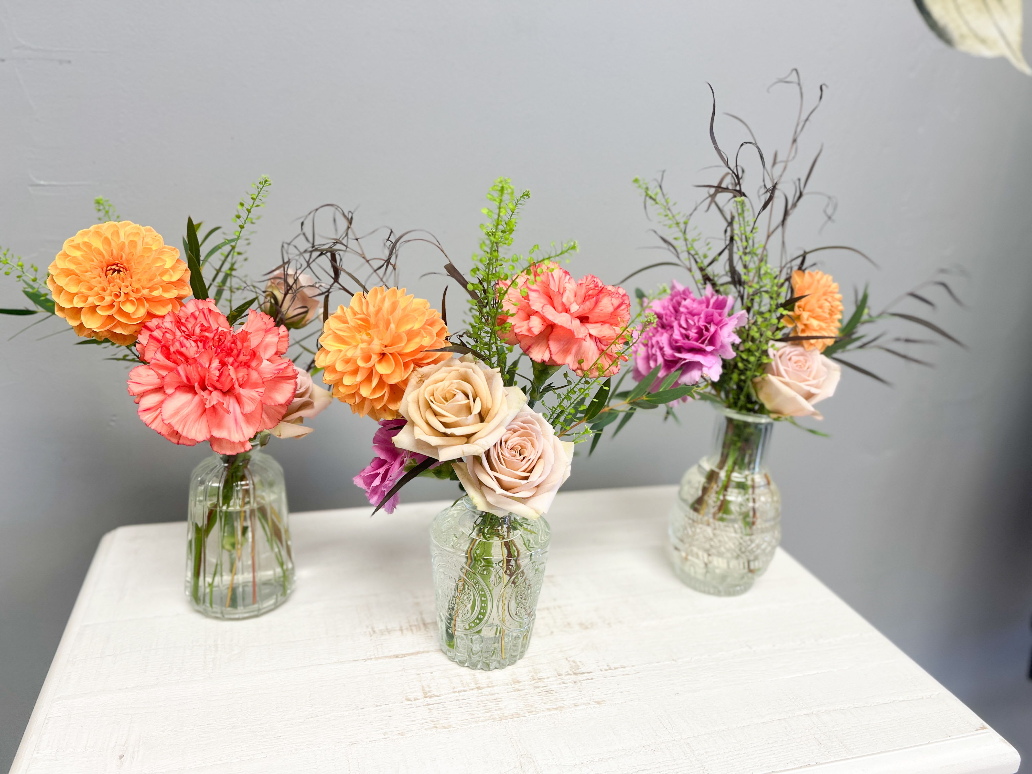 Golden Glow Bud Vases Golden Glow Bud Vases - Brighten any space with our Golden Glow Bud Vase, a petite burst of beauty designed to make a big impression. Each vase features a radiant mix of orange dahlias, coral carnations, soft mauve roses, and textured greenery, thoughtfully arranged in a decorative glass vase. Perfect for gifting, desk décor, or adding a pop of color to your home, these mini arrangements are full of warmth and joy. Features: • Fresh, hand-arranged flowers in a patterned glass bud vase • Vibrant tones of golden yellow, coral, mauve, and green • Long-lasting blooms with easy care • Ideal for gifting, home accents, or small celebrations Care Tip: Change the water every 2–3 days and keep flowers away from direct sunlight to enjoy their beauty longer.