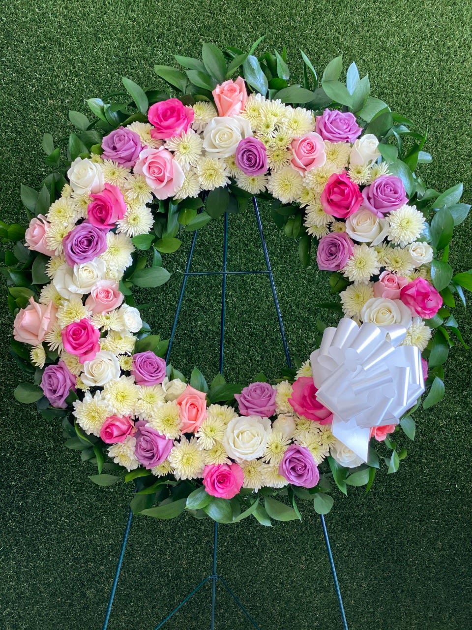 DEEPEST CONDOLENCES - Includes: • Roses • Mums • Greenery • Easel  Celebrate a life with this Pink, purple and white Standing Wreath. The wreath is created by one of our local florist with pink, purple, white roses, white mums, greenery and more. All the flowers are arranged in a wreath shape, a beautiful gift for a funeral. 