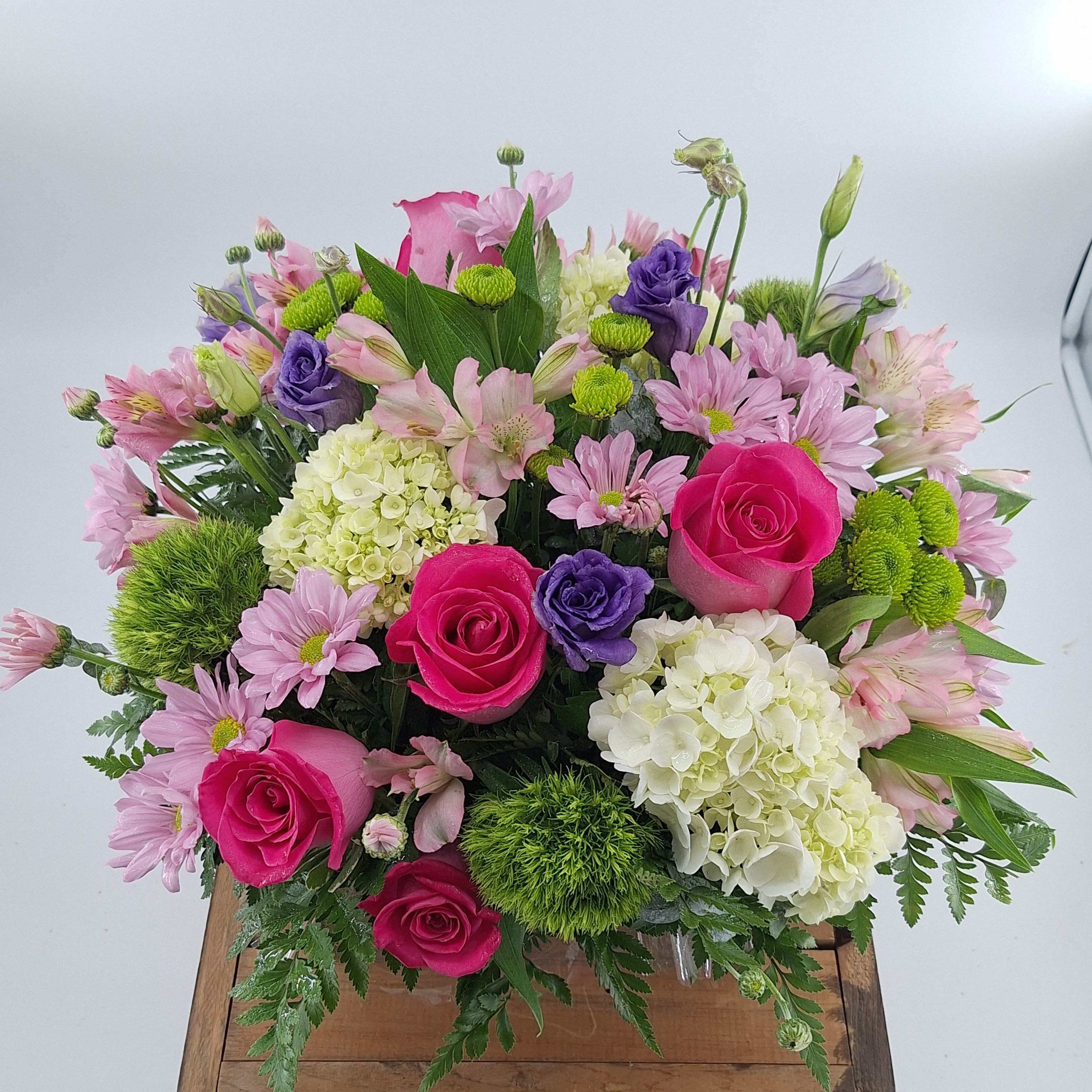 Fascination (DL) - Elevate your space with our modern floral arrangement in a brown 7x7&quot; wooden box. The striking mix of hot pink roses, purple lisianthus, Green trick, mini green hydrangea, pink alstroemeria , green buttons, lavender daisies, and assorted greenery creates a captivating display. This arrangement exudes contemporary elegance, making it the perfect centerpiece to add a touch of sophistication and style to any setting. 