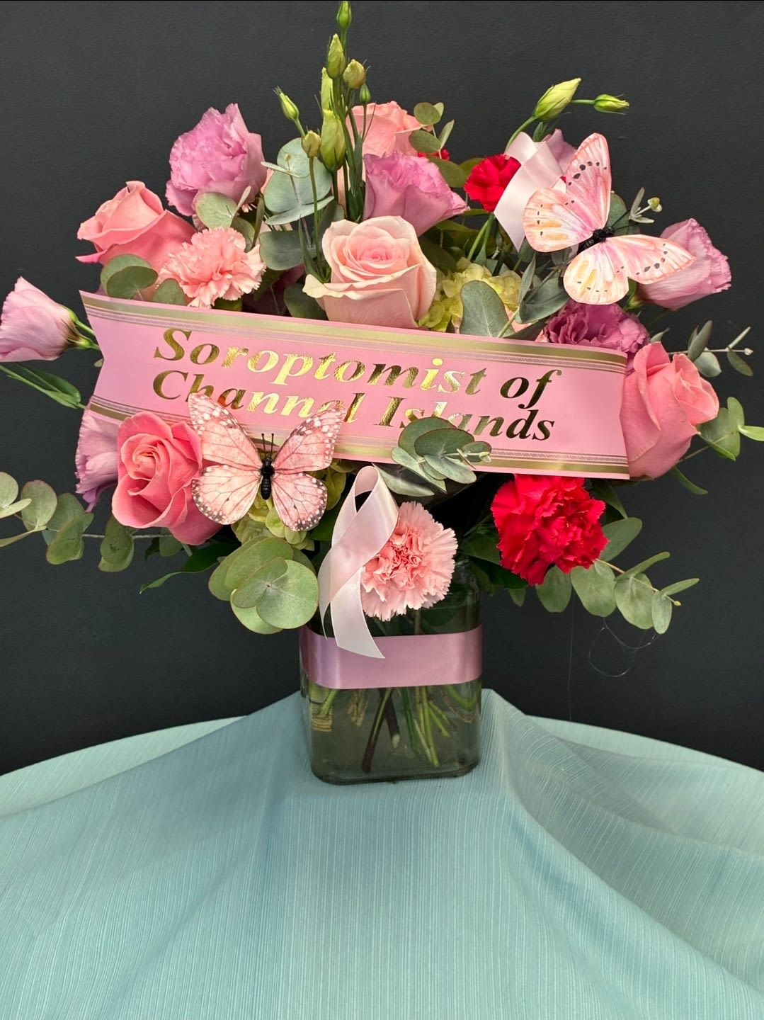 S.C.I Pretty in Pink - **FOR EVERY ARRANGEMENT PURCHASED, 20% WILL BE DONATED TO SOROPTOMIST OF CHANNEL ISLANDS FOR BREAST CANCER AWARENESS***  A radiant celebration of empowerment and elegance, this handcrafted bouquet features lush pink roses, vibrant red and blush carnations, delicate lisianthus, and airy eucalyptus. Nestled among the blooms are whimsical pink butterflies, symbolizing transformation and hope. The arrangement is presented in a clear glass vase wrapped with a soft pink ribbon, resting on a serene seafoam drape. A custom banner in gold and black proudly honors the Soroptimist of Channel Islands, making this piece a heartfelt tribute to women uplifting women.         *****PICTURE DEPICTS THE DELUXE SIZE*** **BANNER NOT INCLUDED, ONLY FOR PORMOTIONAL PURPOSES***