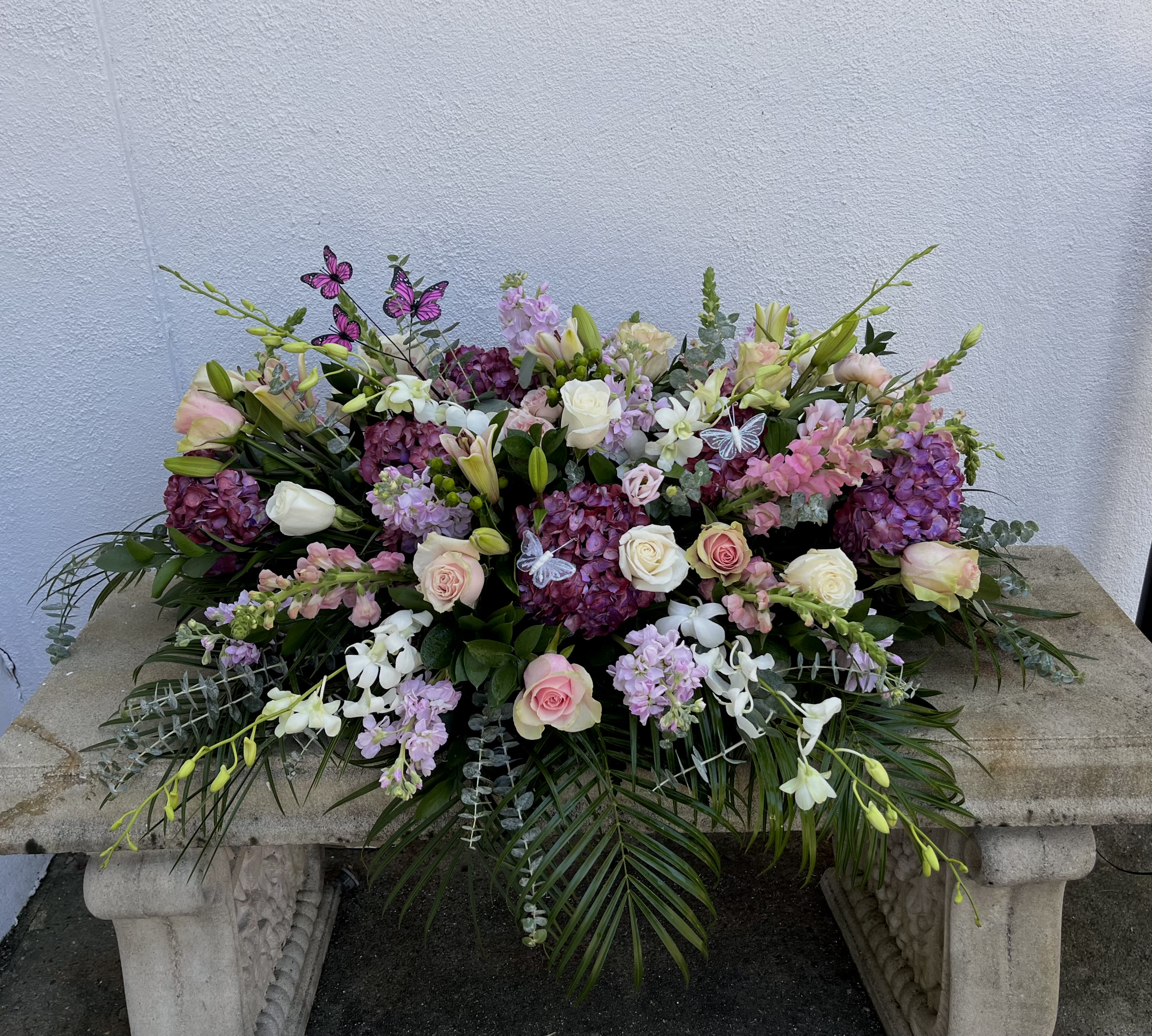 Fluttering Remembrance Casket Spray - Roses, lilies, Hydrangea, stock, snap dragon, hypericum, orchid stems and butterfly accents  come together to create a garden inspired casket spray. Sized to fit both open and closed caskets
