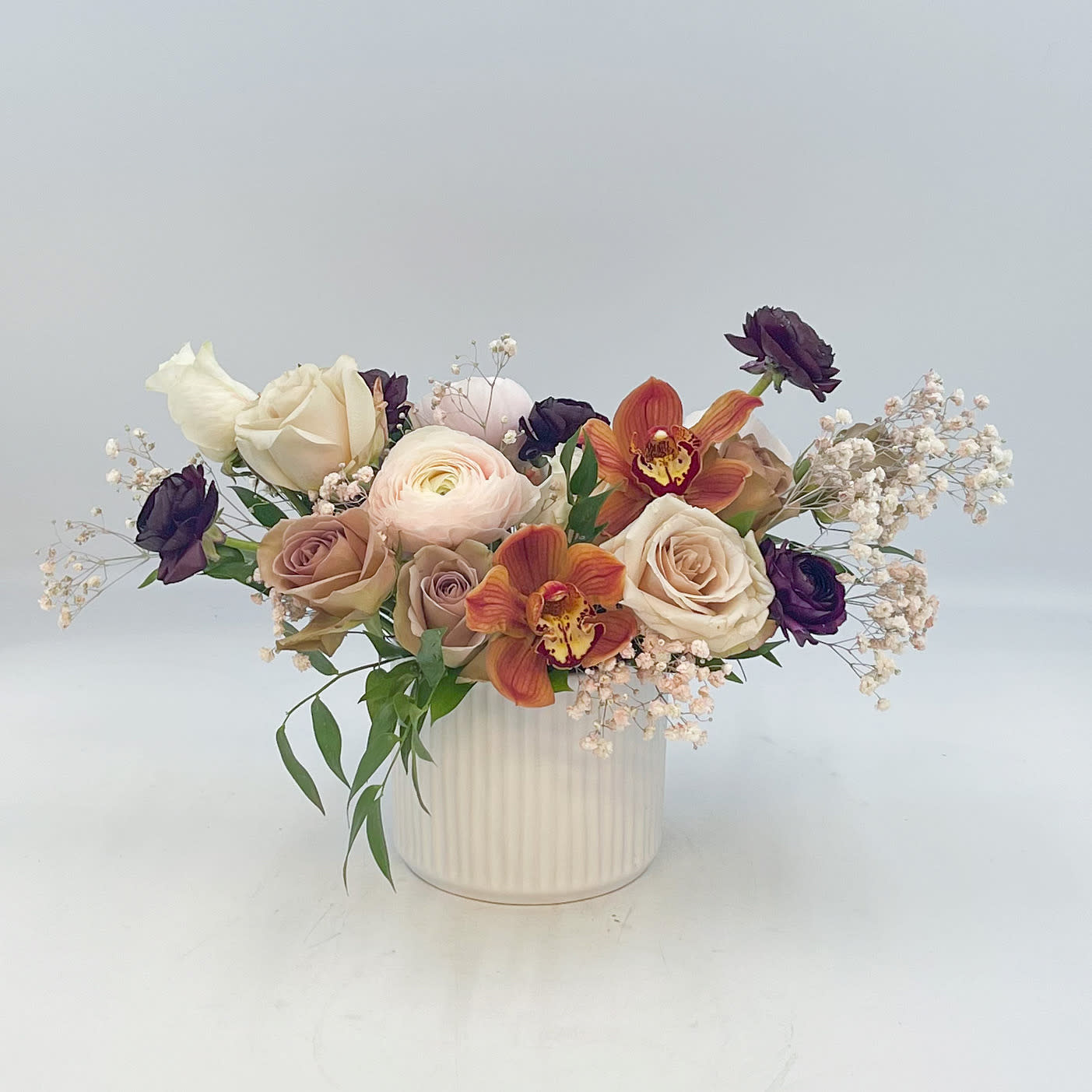 Cheeky - Cheeky is a fun and flirty mix of Chocolate Cymbidium, Blush and Burgundy Ranunculus, Ivory and Sand colored Roses (Vendela, Quicksand and Amensia) with Ruscus and Blush sprayed babies breath arranged in a keepsake Ceramic container and is pictured. 