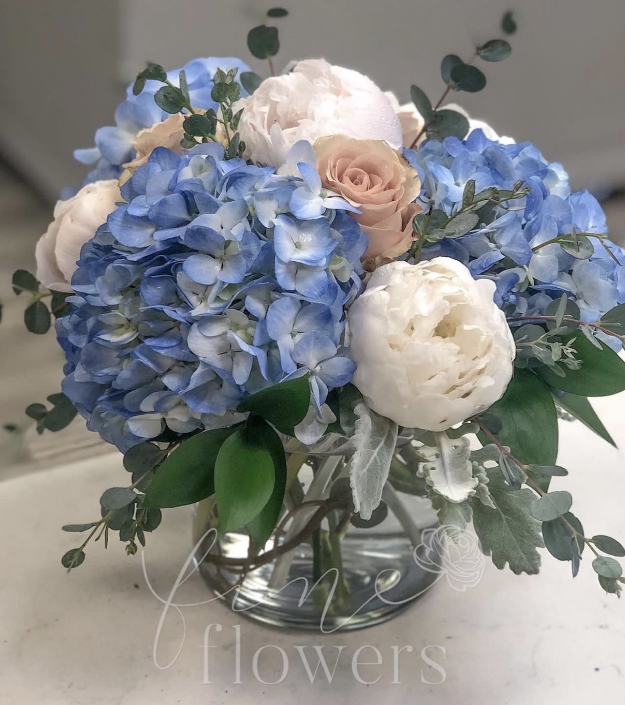 Sandbar - Sandbar is a gorgeous earth toned arrangement for any lover of the beach. Sandbar features Blue Hydrangea, Sand colored Roses, White Peonies, Dusty Miller and a touch of Eucalyptus.  The Medium Sandbar is arranged in a 5 inch clear glass cylinder and the Large Sandbar is arranged in a 6 inch clear glass cylinder.