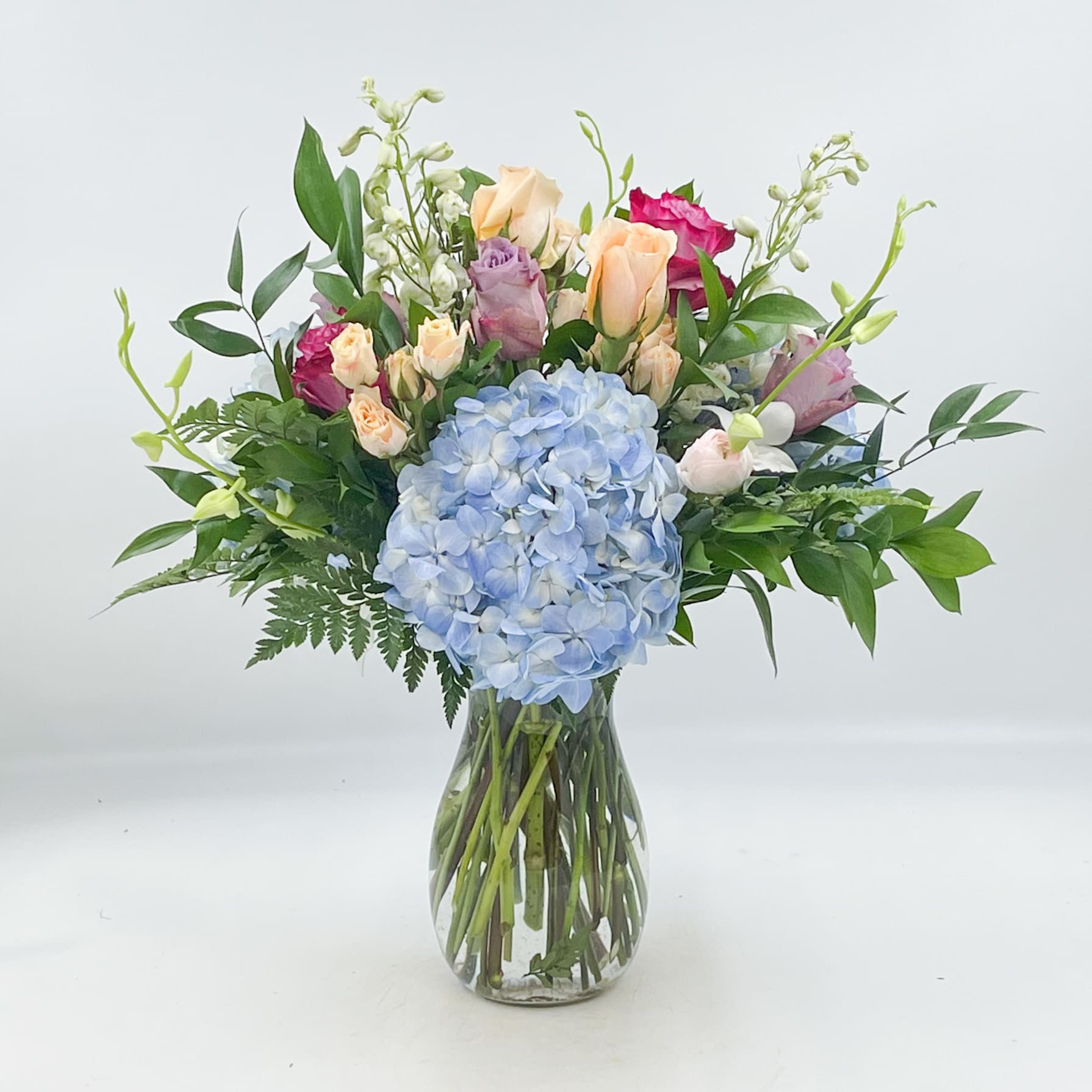 Monet - Monet is a dreamy and colorful pastel arrangement. Monet is truly a work of art, with its Blue Premium Hydrangea, Peach Roses, Deep Purple Roses, Lavender Roses, Pink Ranunculus, Peach Spray Roses, White Delphinium Orchid Stems and Mixed Ruscus and Ferns.