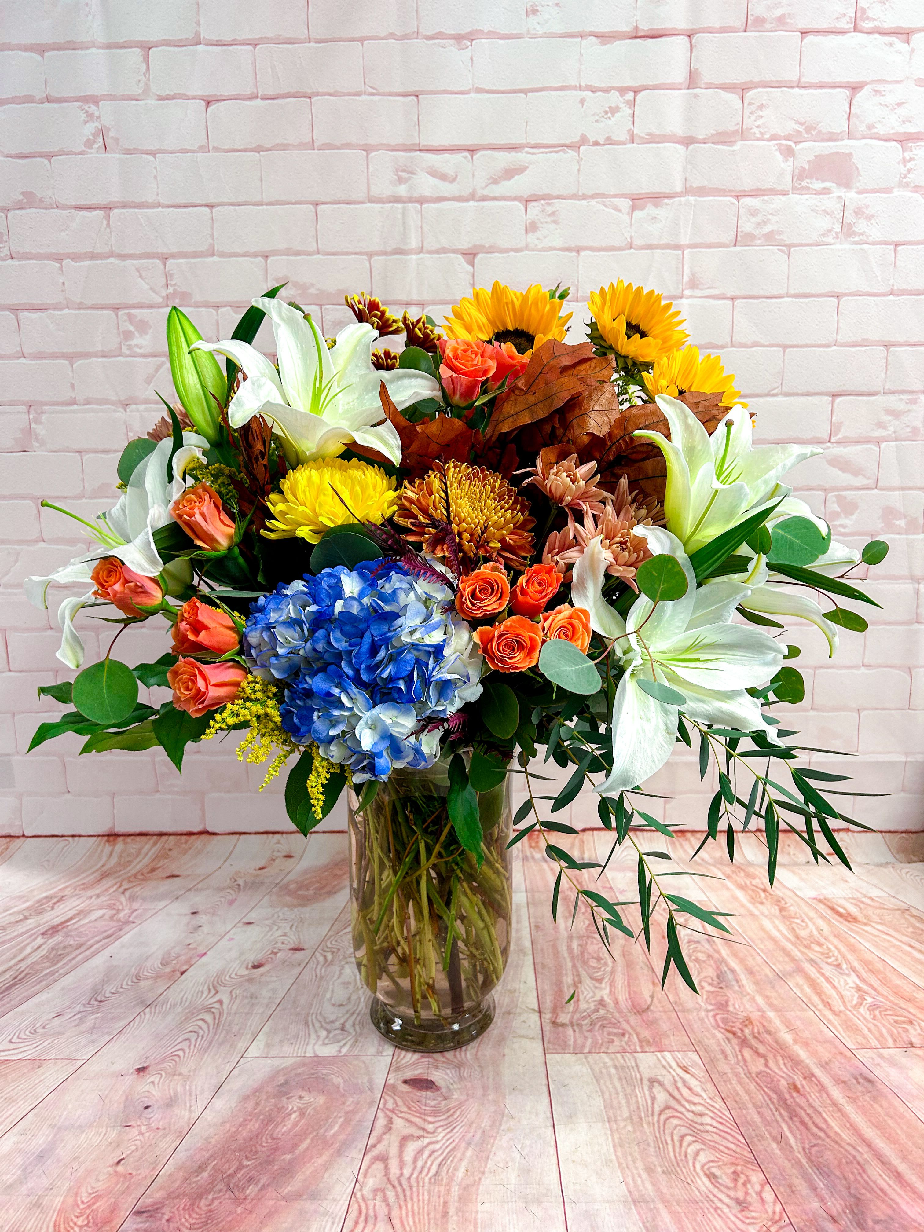 Autumn Radiance Bouquet - Celebrate the beauty of the season with our Autumn Radiance Bouquet — a stunning display of nature’s most vibrant hues. This arrangement features a rich blend of golden sunflowers, white lilies, orange spray roses, and blue hydrangeas, accented with textured autumn leaves and lush greenery.  Perfect for brightening a home, sending thanks, or celebrating a special occasion, this arrangement brings the warmth of fall indoors. Hand-designed in a clear glass vase, it’s a breathtaking centerpiece that captures the essence of autumn’s charm and color.