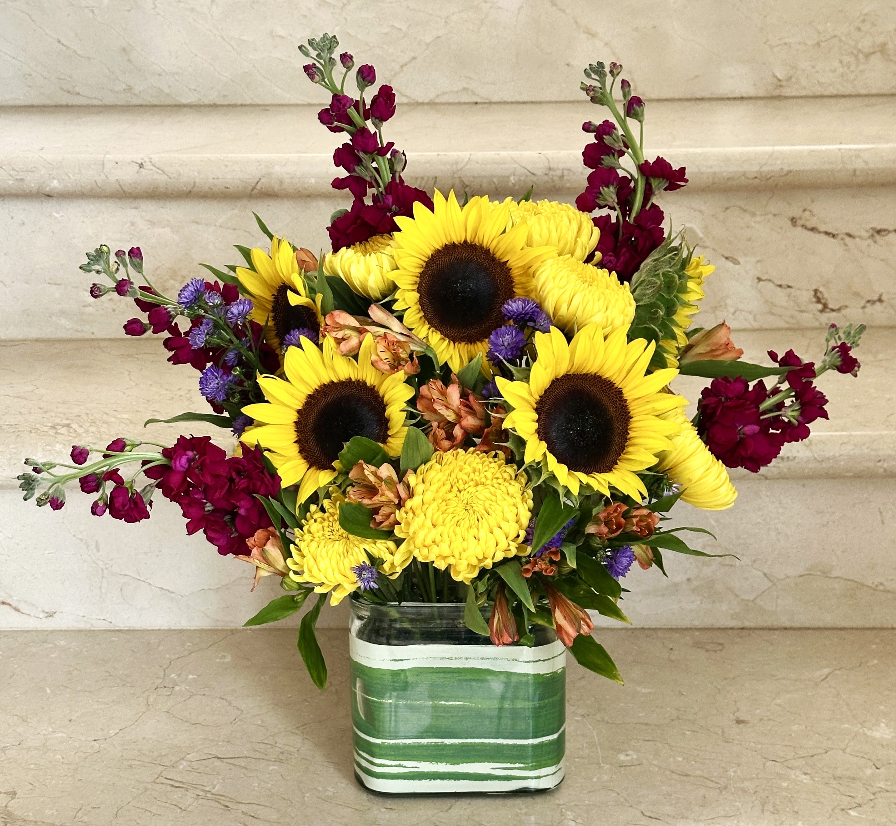 Sunset Dreams - A cheerful mix of sunflowers, burgundy snapdragons, yellow mums, and other seasonal flowers in a stylish green glass vase. Perfect for birthdays, get well wishes, or just because. 