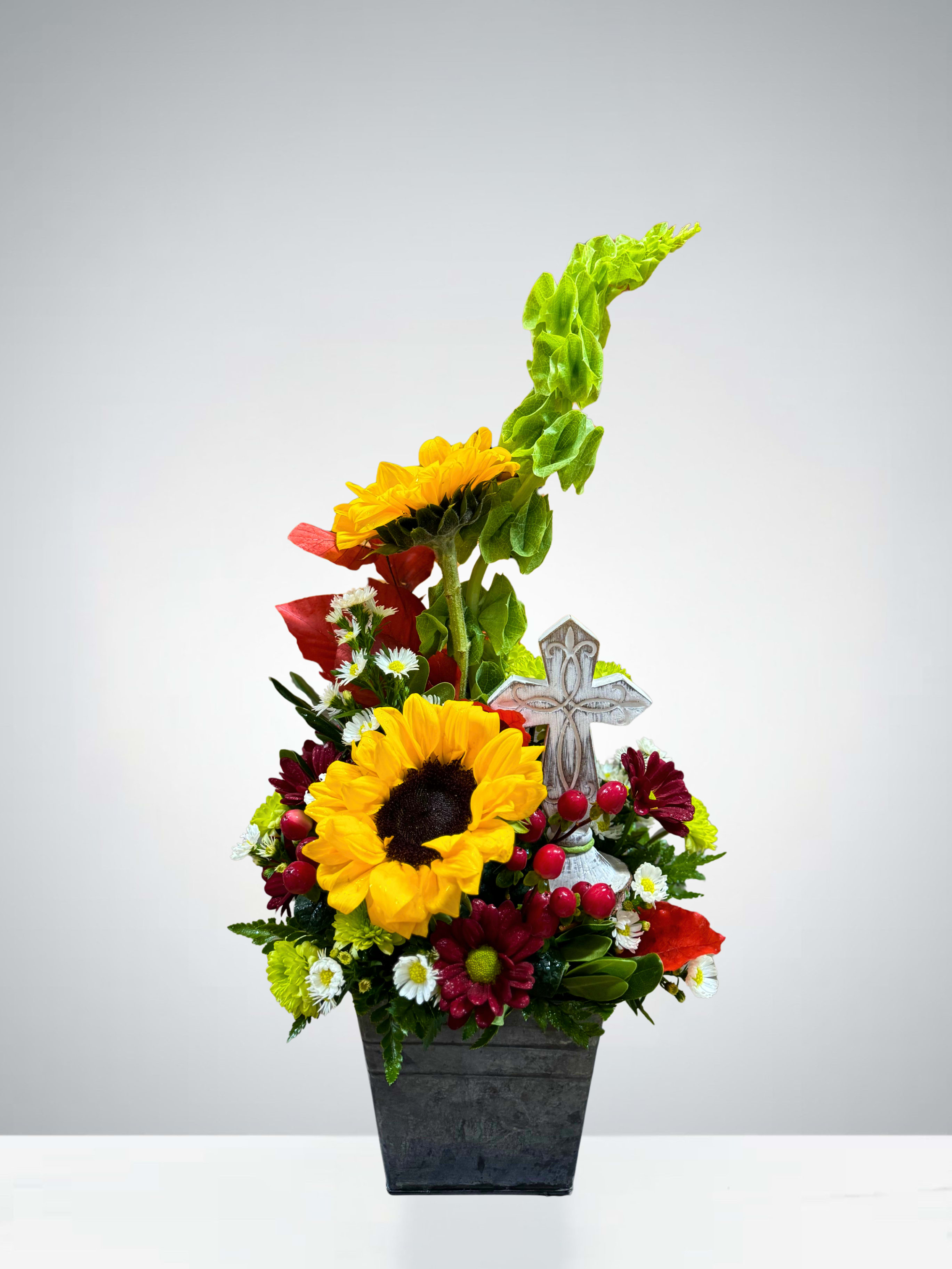 Faithful Sunshine - Bright and uplifting, this arrangement brings warmth and comfort through cheerful sunflowers, rich red berries, bell of ireland, white monte, red daises, and fresh greenery. Accented with a decorative cross, it serves as a beautiful reminder of faith, hope, and love. Perfect for expressing sympathy, offering encouragement, or celebrating a life well-lived.  (Flower varieties and colors may vary based on availability.)