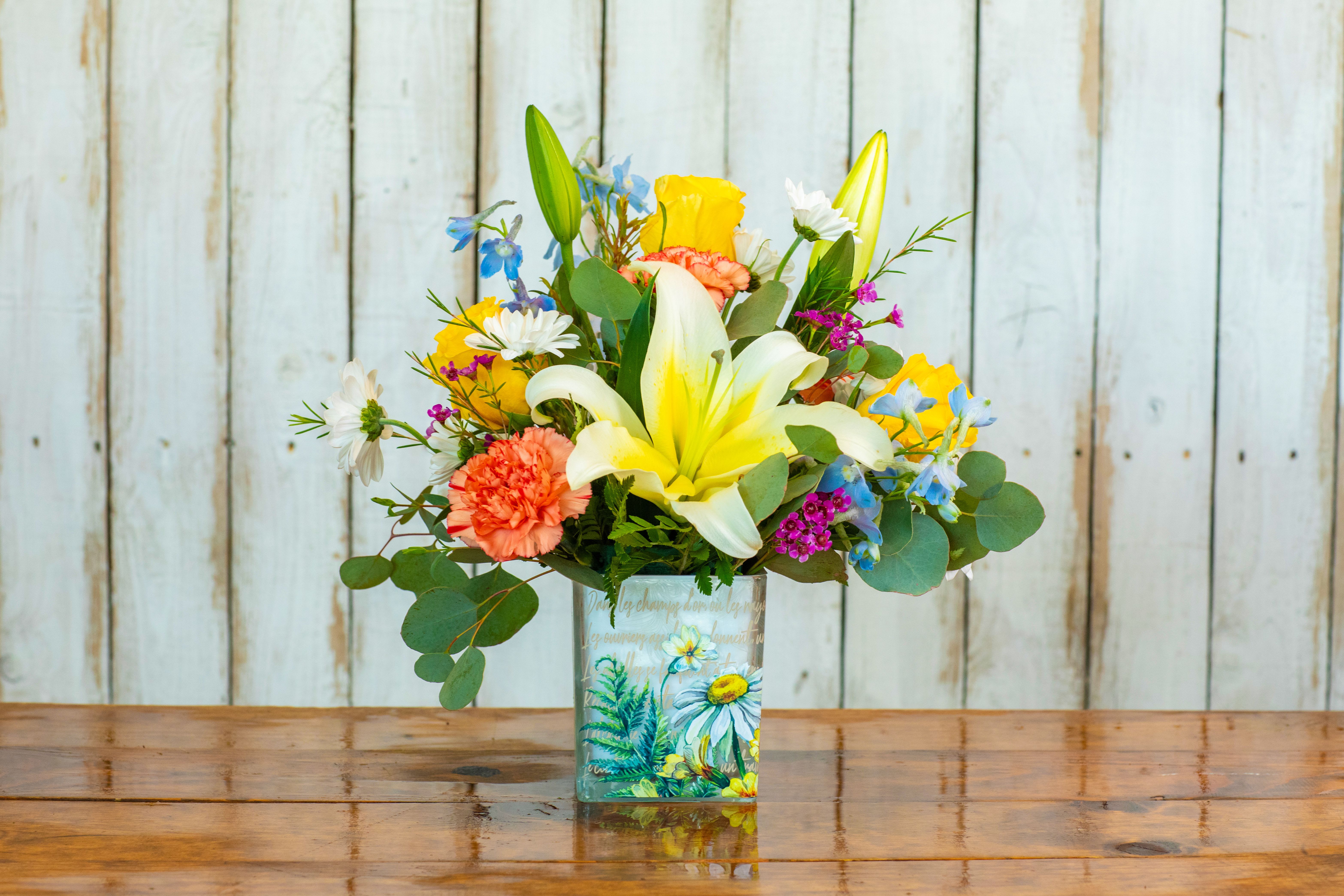 Golden Meadow Bouquet Golden Meadow Bouquet - Brighten any day with the Golden Meadow Bouquet, thoughtfully arranged by your trusted Rancho Cucamonga Florist, Tommy Austin Florist. This cheerful and vibrant bouquet evokes the joyful feeling of a sun-drenched meadow. It features a delightful mix of sunny yellow lilies taking center stage, surrounded by cheerful yellow roses, delicate peach carnations, and a scattering of charming white daisies. Playful accents of blue delphinium and other colorful filler flowers add depth and texture, all brought together with fresh eucalyptus greenery. The arrangement is presented in a unique glass vase adorned with a picturesque floral illustration, making it a perfect gift for birthdays, thank yous, or simply to bring a touch of outdoor beauty indoors. Let Tommy Austin Florist deliver a smile with the "Golden Meadow" throughout Rancho Cucamonga and neighboring areas.