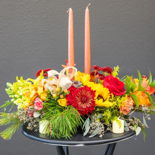 Gather 'Round Gather 'Round - This beautiful fall center piece arrangement is sure to have everyone clamoring around the dinner table to share stories of what they are grateful for. Gather "round features yellow sunflower, red cremone, roses, and snap dragons, along with other accents and fall embellishments. This item will include 2 candles.