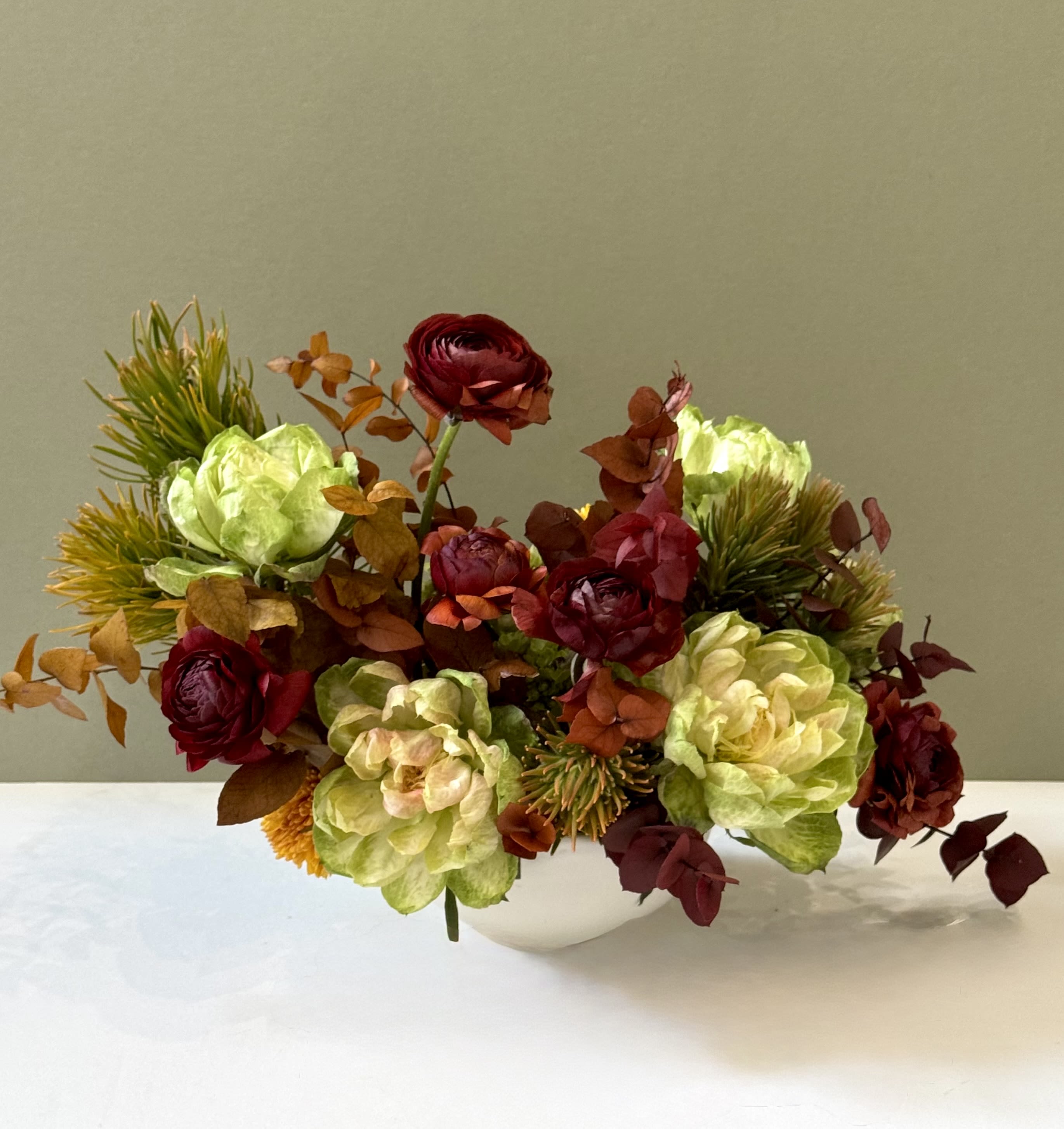Amber Groove - A striking fall composition blending deep burgundy ranunculus, warm amber foliage, and soft green blooms. Textural and modern, this arrangement captures the glow of autumn forests — bold yet refined, perfect for bringing seasonal warmth to any space.