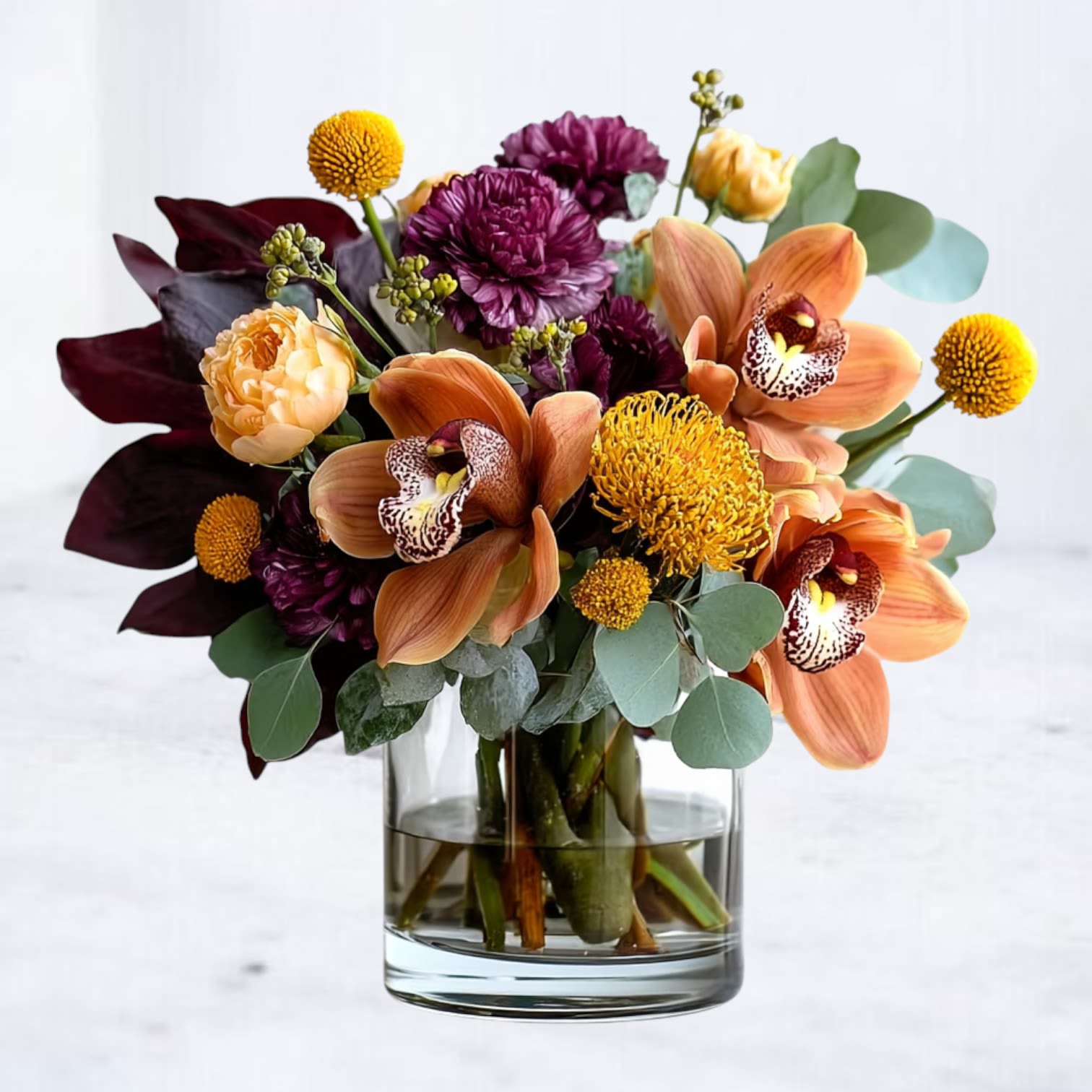 Kindled Earth Kindled Earth - A sculptural study in autumn’s warmth and depth, Kindled Earth blends the grounded tones of the season with modern refinement. Burnt amber orchids, golden pincushion protea, and plum chrysanthemums meet soft peach ranunculus and eucalyptus in a lush, artfully layered arrangement. Each bloom seems lit from within, echoing the quiet glow of firelight and the richness of freshly turned soil. Designed in a sleek glass cylinder, this bouquet bridges the space between organic and opulent — perfect for gifting, seasonal centerpieces, or as an expressive statement of gratitude.