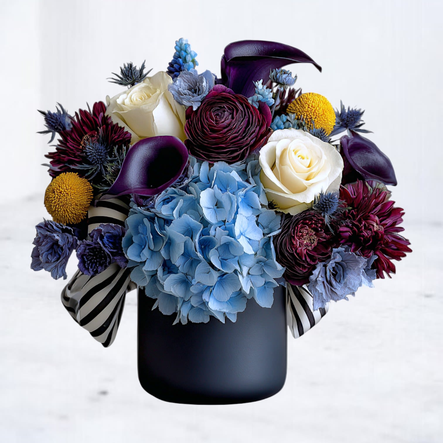 Vanta Veil Vanta Veil - Bold, enigmatic, and unapologetically chic — Vanta Veil captures the allure of darkness wrapped in elegance. This striking arrangement layers deep plum calla lilies, blue hydrangea, creamy roses, and textured blooms of gold and maroon, all styled within a black vase. Like shadows meeting moonlight, every petal and hue plays with contrast — soft ivory against midnight tones, rich textures framed by sleek modern design. Finished with a subtle striped accent, this bouquet feels at once fashion-forward and timeless, an embodiment of beauty with an edge. Perfect for gifting when words fall short — for anniversaries, creative souls, or those drawn to art, mystery, and depth.