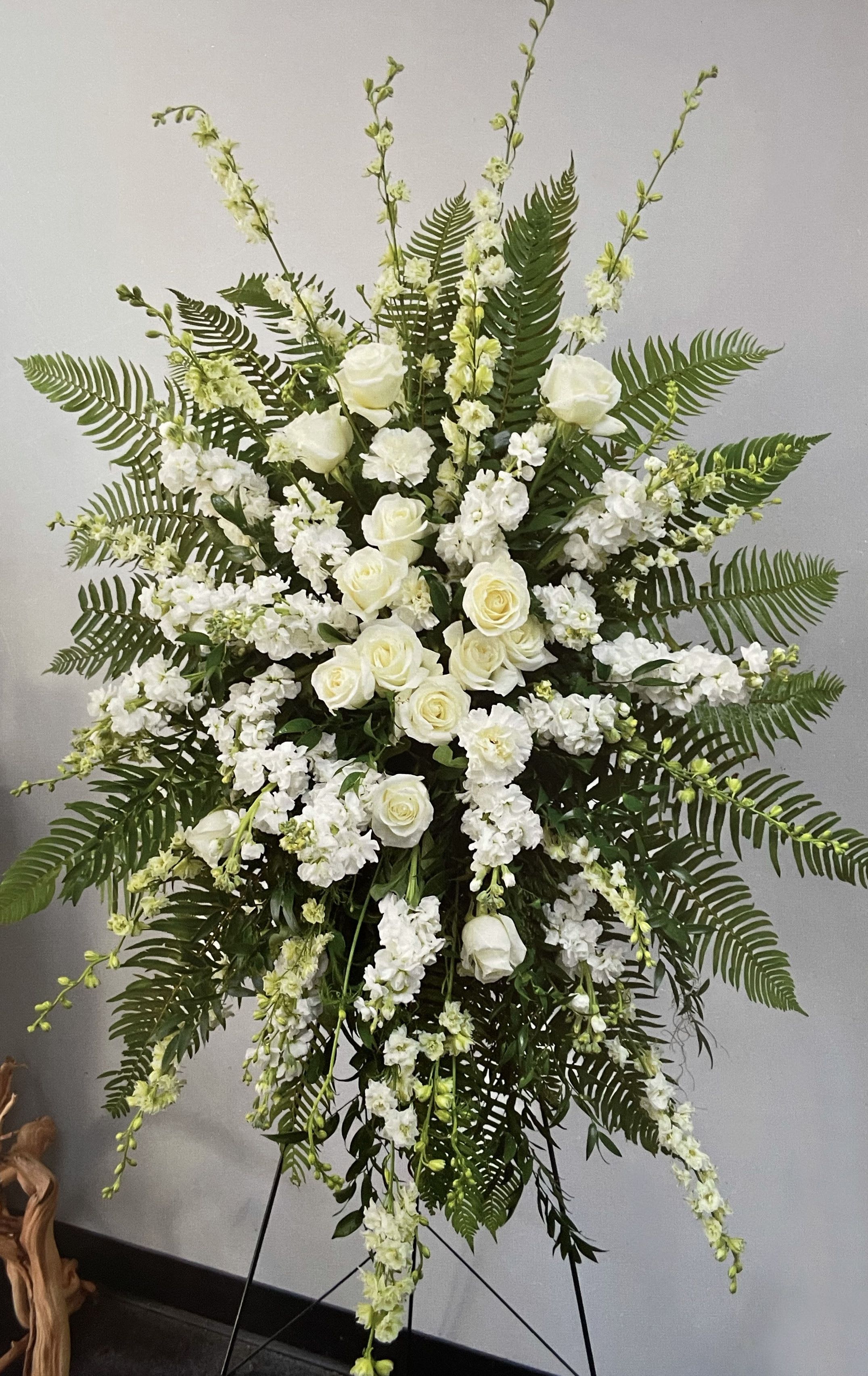 Easel Spray ES-09 Easel Spray ES-09 - Simple and graceful, this white and green standing spray offers a timeless expression of peace and reverence. Creamy white roses, larkspur, and stock are artfully arranged among fresh ferns and soft greenery, creating a full and elegant shape. Displayed on an easel (stand included), this design is a beautiful way to honor a loved one with purity, warmth, and quiet dignity.