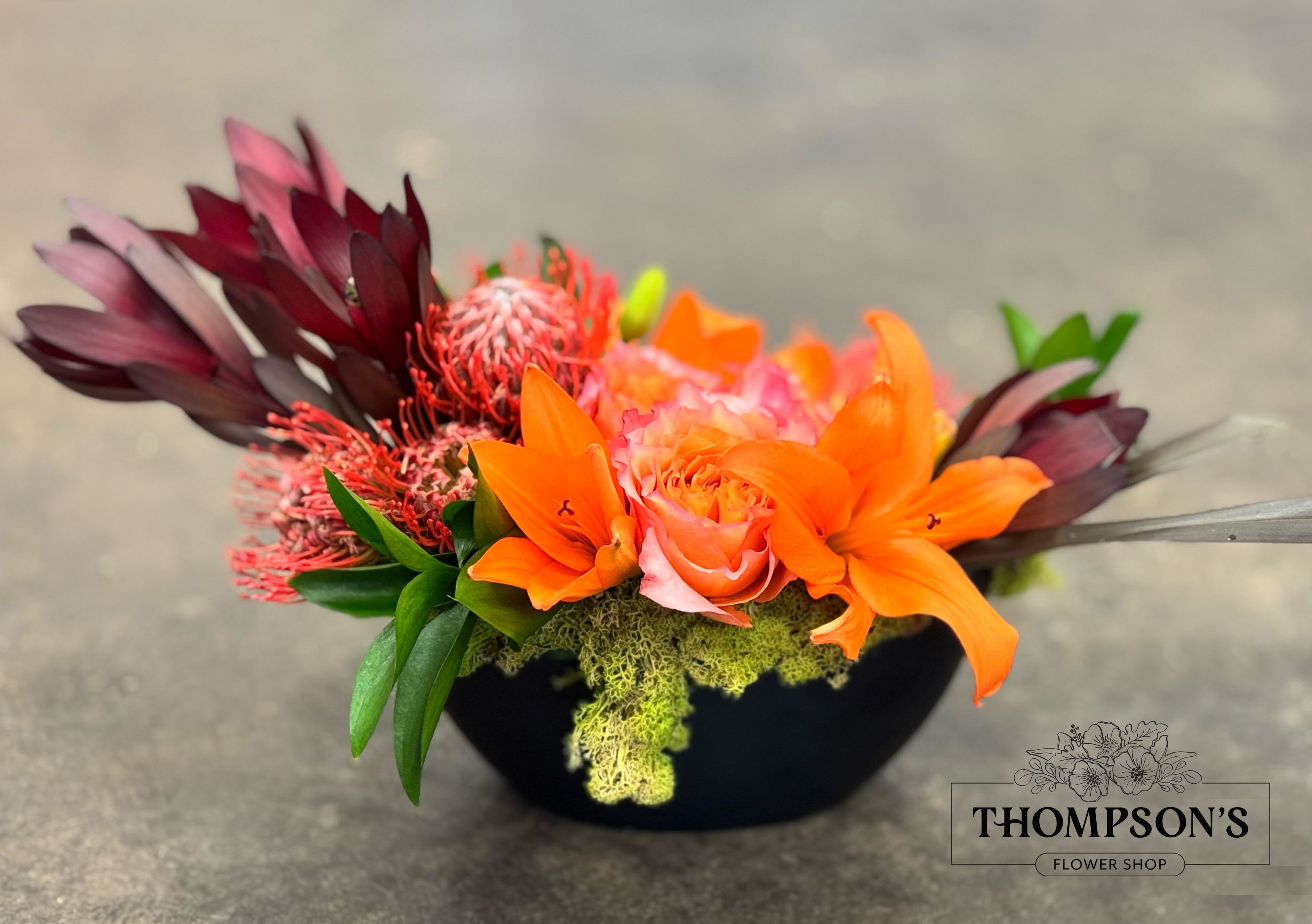Vermillion - Vermillion captures the warmth of curated color, blending fiery orange tones with deep burgundy accents in a refined, modern form. Designed in a low, contemporary ceramic vase, this arrangement celebrates movement, texture, and contrast without overpowering the space. Tropical blooms and premium florals are styled with an artist’s touch,  ideal for a one-of-a-kind gift.