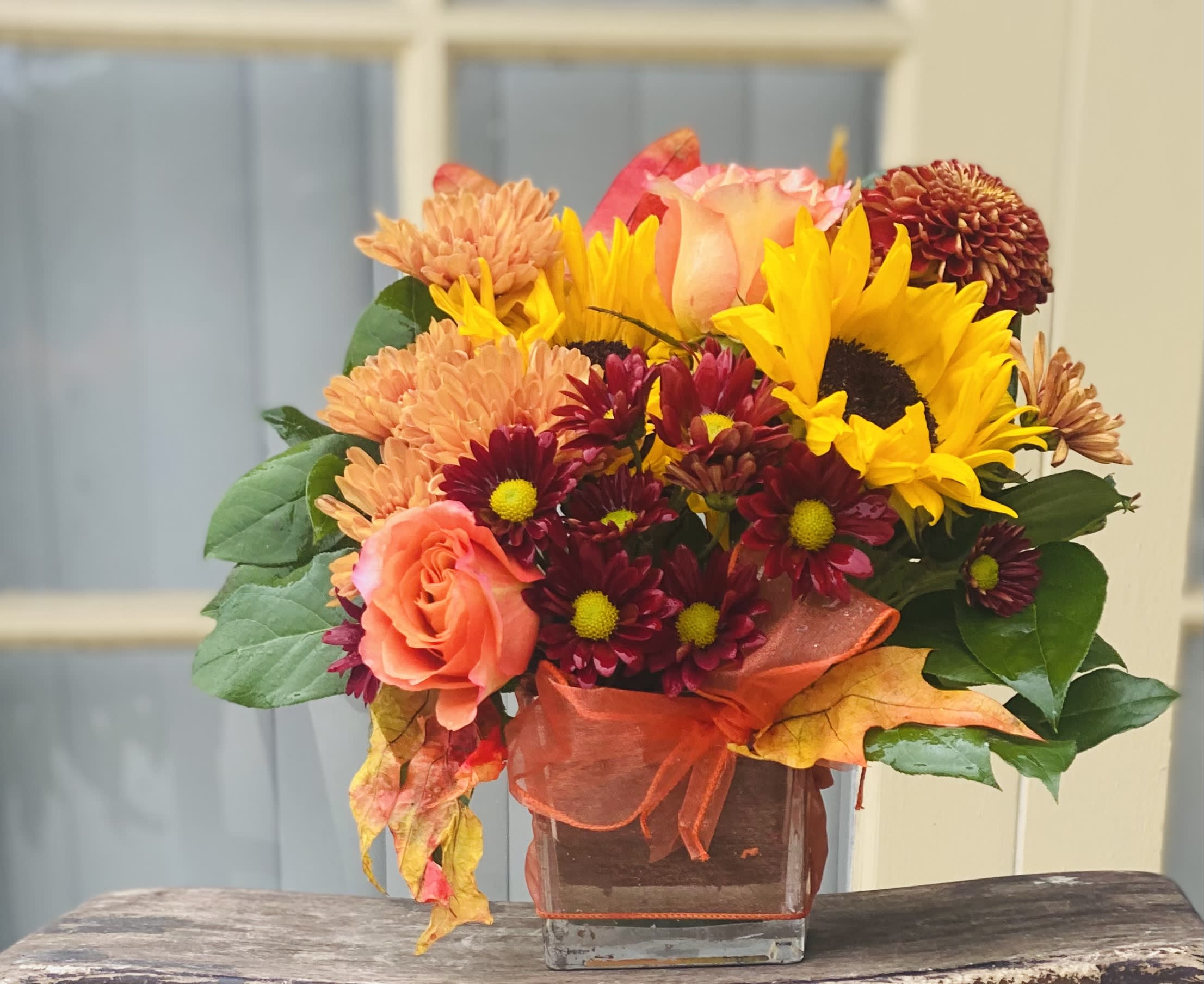 Orange Roses &amp; Sunflowers Mix Autumn Gift  - This fall-themed gift showcases the rich colors of autumn. It includes eye-catching orange roses, evoking the season's foliage, paired with sunny yellow sunflowers. Soft orange and burgundy blossoms provide a warm and welcoming feel. This creation makes a wonderful present to cheer someone up, perfect for celebrations or just adding a smile.
