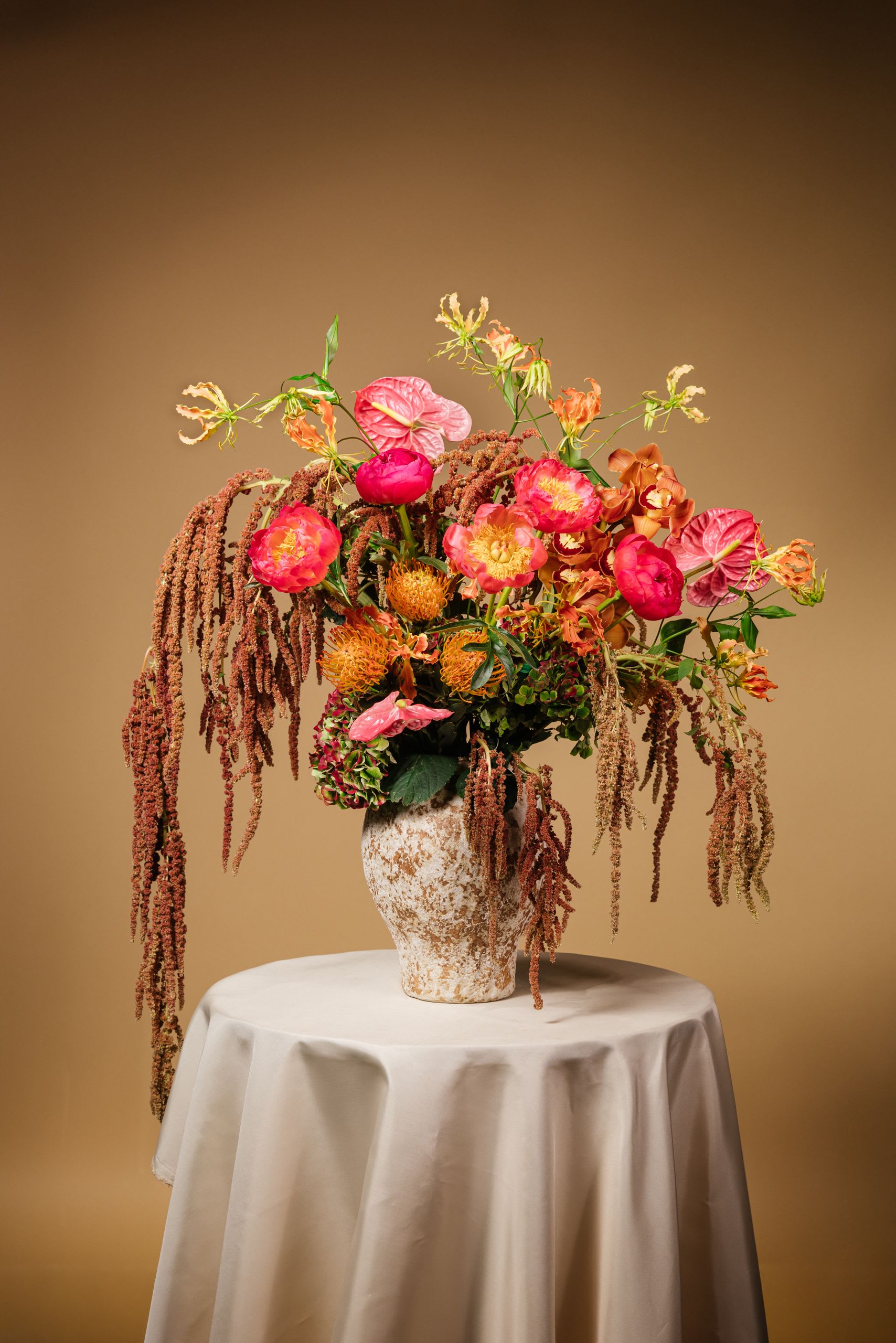 Sunset Reverie Arrangement with Vase - The Sunset Reverie Arrangement captures the warmth and radiance of a tropical evening sky. A luxurious blend of coral peonies, orange pincushion proteas, pink anthuriums, gloriosa lilies, orchids, and amaranthus cascades gracefully from a textured ceramic vase. The vibrant hues of coral, gold, and deep amber create a striking visual harmony—perfect for modern weddings, island-inspired events, or as a statement centerpiece that brings the energy of a Hawaiian sunset indoors.