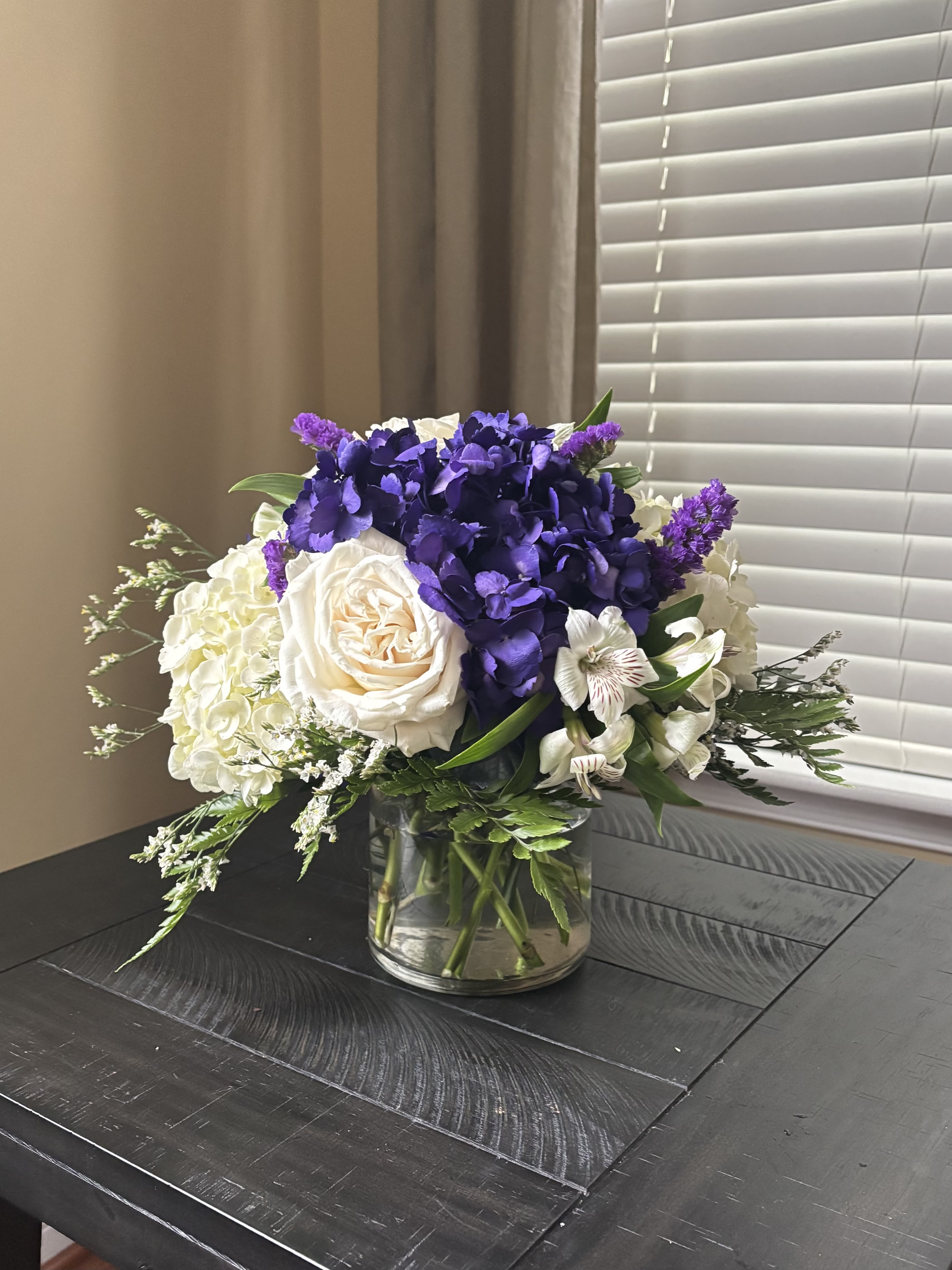 Purple and Cream - Soft, elegant, and timeless — this arrangement of purple hydrangeas, white hydrangeas, and white roses brings a touch of refined beauty to any space. The rich purple tones add depth and luxury, while the crisp white blooms create a clean, classic balance.  Presented in a stylish small glass vase, this design is perfect for coffee tables, desks, or intimate gatherings. Each bloom is carefully selected and artfully arranged to highlight the natural harmony of color and texture.  Includes: Purple hydrangeas White hydrangeas White roses Seasonal greenery Small clear glass vase  Graceful and fresh, this arrangement captures the perfect blend of sophistication and serenity — ideal for gifting or adding a touch of elegance to your home.