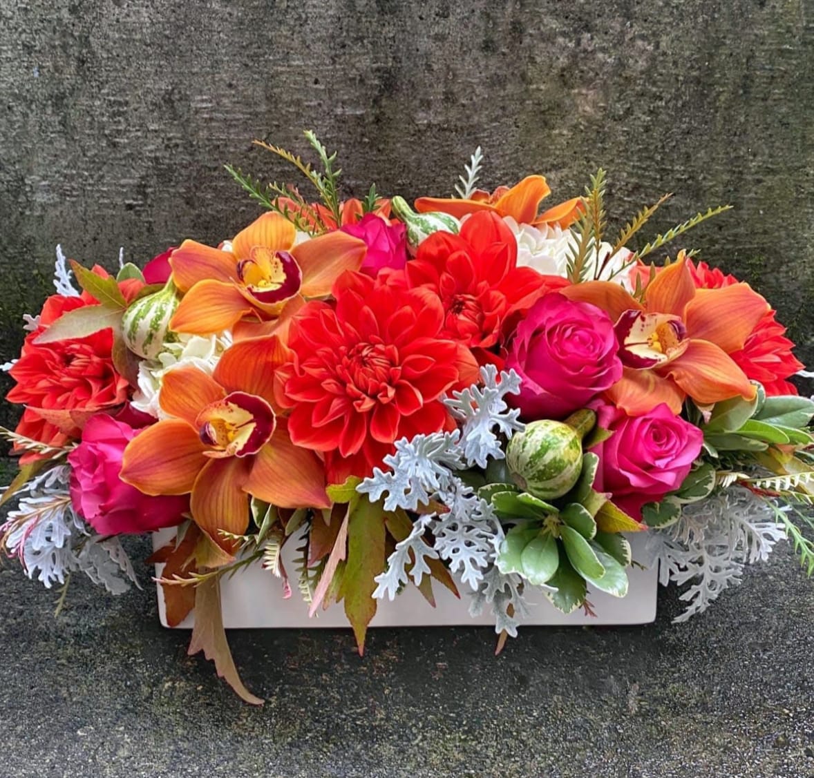 Fall Mix Long and Low Centerpiece - Fresh flowers are the secret to a well-dressed and welcoming table. Studies have shown that flowers are a natural mood elevator and a centerpiece from Fiori Floral Design is sure to be a conversation piece. You may not be able to celebrate in person with those you love this year but you can let them know that you are with them in spirit and put the finishing touch on their table with a gift of flowers.  Our Fall Mix Collection are Designer's Choice arrangements that celebrate the colors and flowers of autumn. We accent them with fall leaves such as weet gum and often add fruits or berries such as rosehips or Tennessee spinning gourds from the cutting garden. Every arrangement will be different depending our weekly color palette, what is in season in the cutting garden and what the flower farms bring to the local flower co-op.  Out Long and Low Centerpiece is perfectly scaled to allow easy conversation without blocking anyone's view. You are welcome to call us with special requests or let us know your first and second choices in the 'Special Requests' box when you place your order online..  We can also match your linen colors on request; just give us a call or leave a note under &quot;Special Instructions&quot; when you place your order.   Approximate dimensions: 10&quot; tall by 7&quot; wide and 15&quot; long.