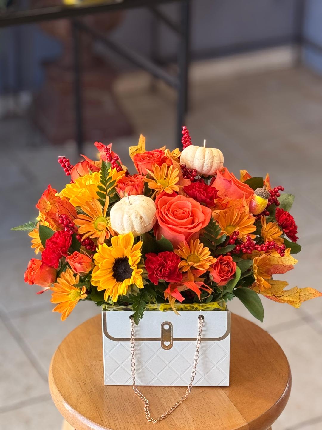 The Harvest Purse - Bursting with rich autumn colors, this white purse overflows with sunflowers, roses, mums, and festive accents like mini pumpkins and fall foliage. Designed to capture the cozy beauty of the season, it makes a perfect centerpiece or a thoughtful gift to celebrate autumn gatherings.  (Flower may vary)