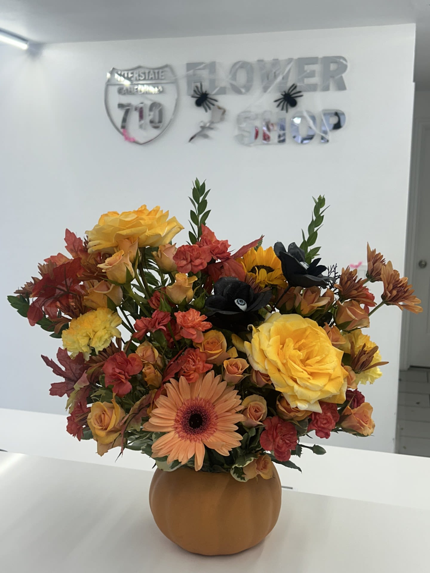 Harvest Whispers - Embrace the cozy magic of fall with this radiant pumpkin arrangement. Bursting with sunlit roses, peach gerbera daisies, and rich autumn foliage, it’s grounded by dramatic black blooms for a bold seasonal twist. Perfect for Halloween, Thanksgiving, or simply celebrating autumn’s golden touch. (If some flowers and/or the vase are not available there will be minor substitutions.)