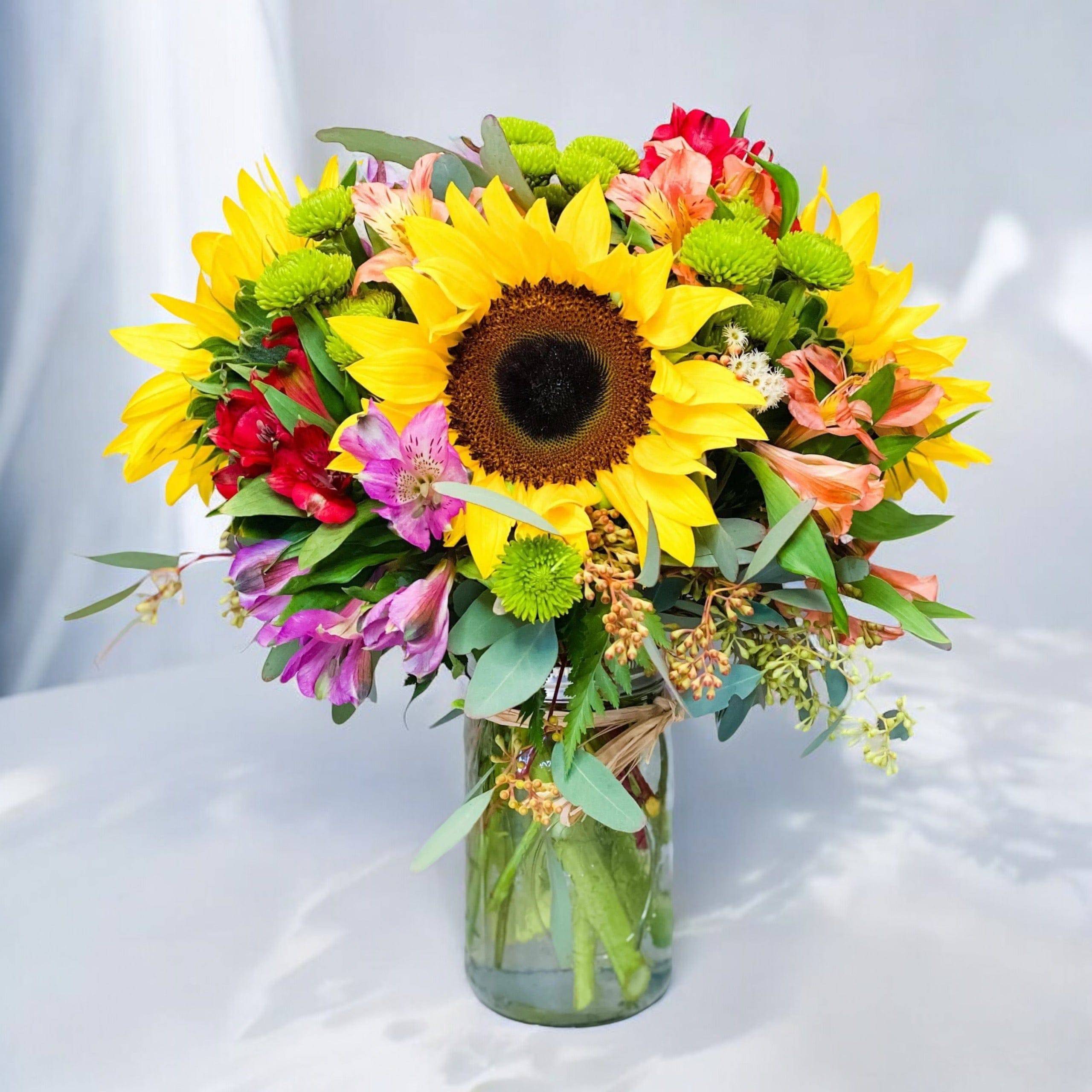 Field Of Flowers Mason Jar Field Of Flowers Mason Jar - If you've ever sauntered through a field of beautiful flowers, you know what a treat it is for the senses and that is what your recipient will come to enjoy with our Field of Flowers Mason Jar. This vividly colorful arrangement captures the essence of a flower field with magnificent sunflowers, alstroemeria and poms set in a stylish mason jar with a natural raffia bow. Treat yourself or surprise a loved one with this delightful mason jar flower arrangement today. Measures 10"H X 8"L. Due to the ongoing supply chain challenges, there is currently a Mason Jar shortage. Therefore we may have to substitute the mason jar with a similar clear glass vase when supply is unavailable