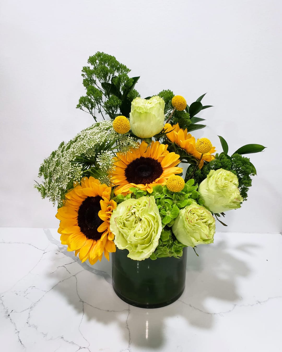 Sunlight - This arrangement has sunflowers, green roses, craspedias, a hydrangea, queens lace, tracelium in a clear glass cylinder vase.