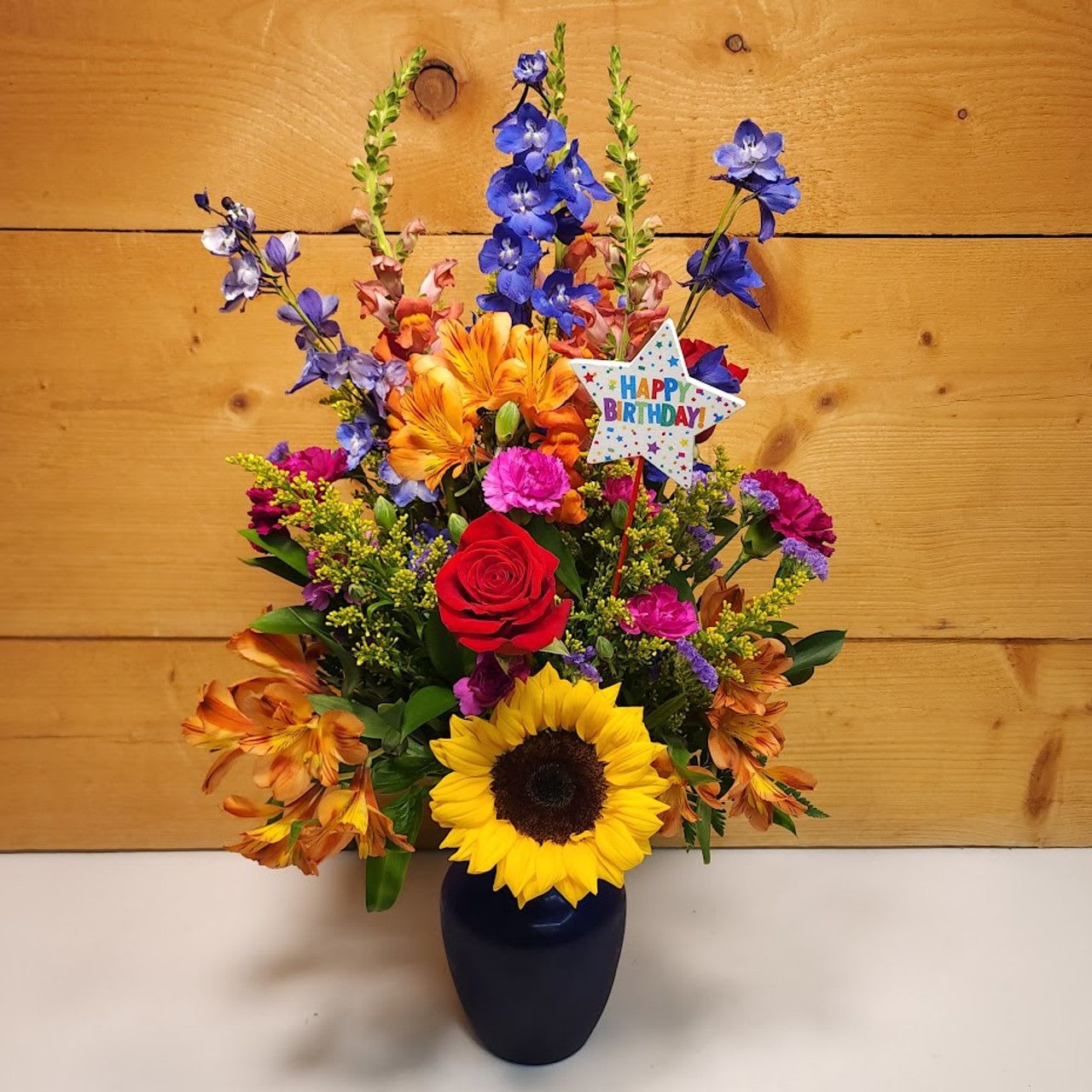 Birthday Radiance Birthday Radiance - Experience the warmth and joy of a perfect day with the "Birthday Radiance" floral arrangement, a dazzling bouquet that radiates cheer and elegance. Nestled in a striking blue vase, this arrangement showcases the sunflowers' golden glow, symbolizing happiness and positivity. Delphinium adds a touch of grace with its tall, enchanting spikes of blue, perfectly complementing the vase. Snapdragons bring vertical interest and a playful spirit with their vibrant colors and unique shape. Mini carnations and carnations offer a soft, lush texture in varied shades, while Alstroemeria adds a delicate, exotic flair. Solidago introduces a burst of golden brightness, and statice provides a subtle, airy texture that enhances the bouquet's overall charm. Together, these flowers create a harmonious and uplifting display, perfect for celebrating the special occasion with a touch of birthday brilliance.
