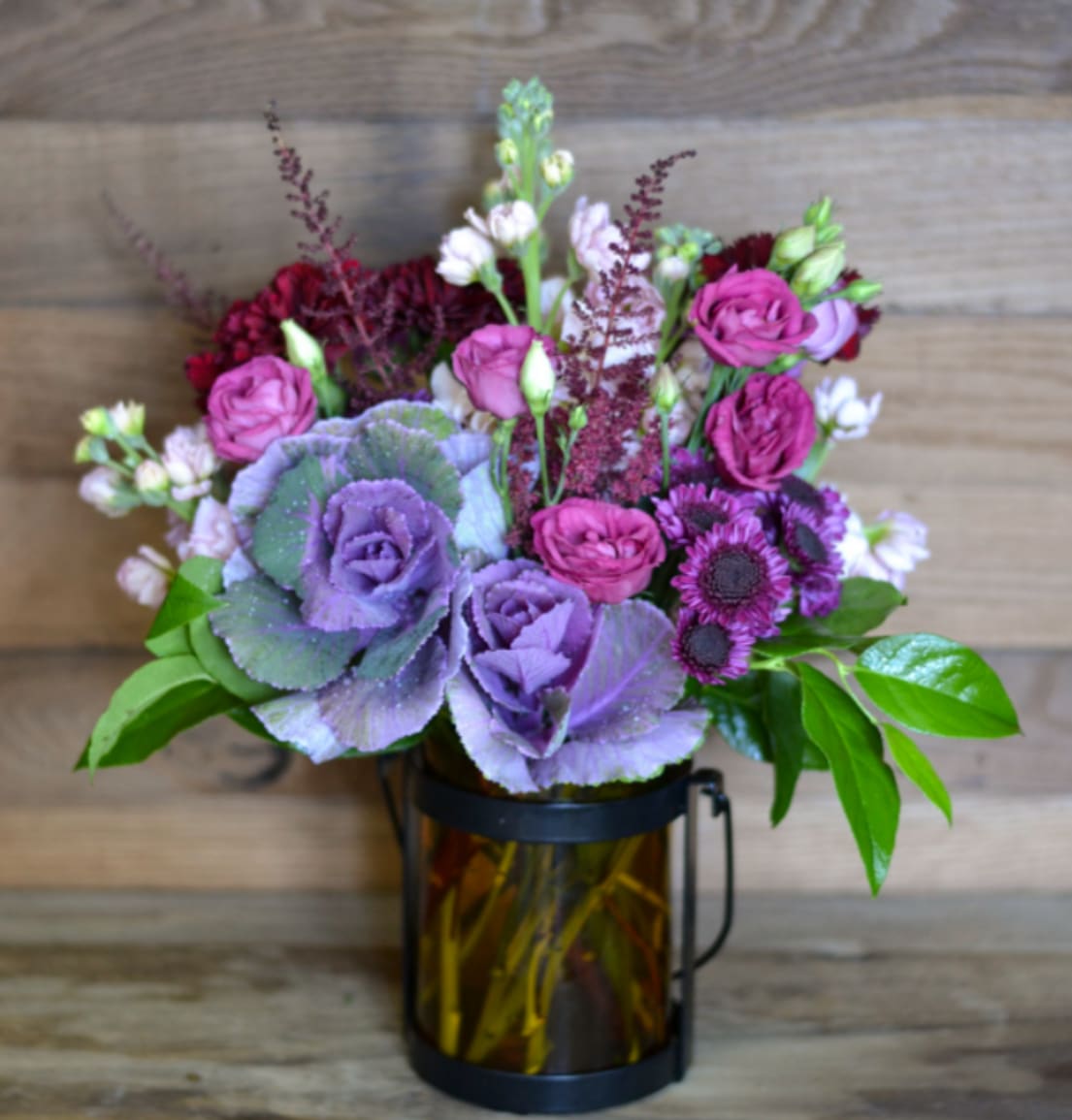 Midnight Sky Bouquet - Light up the night with this beautiful Midnight Sky Bouquet, designed in a petite Amber Lantern that includes a candle for future uses. Our standard bouquet is designed with Ornamental Kale, Rose Lisianthus, Purple Buttons, Pink Stock, and Burgundy Carnations with an upgrade including full sized Roses in addition to the standard design. Our Autumn collection is perfect for nearly any occasion. 
