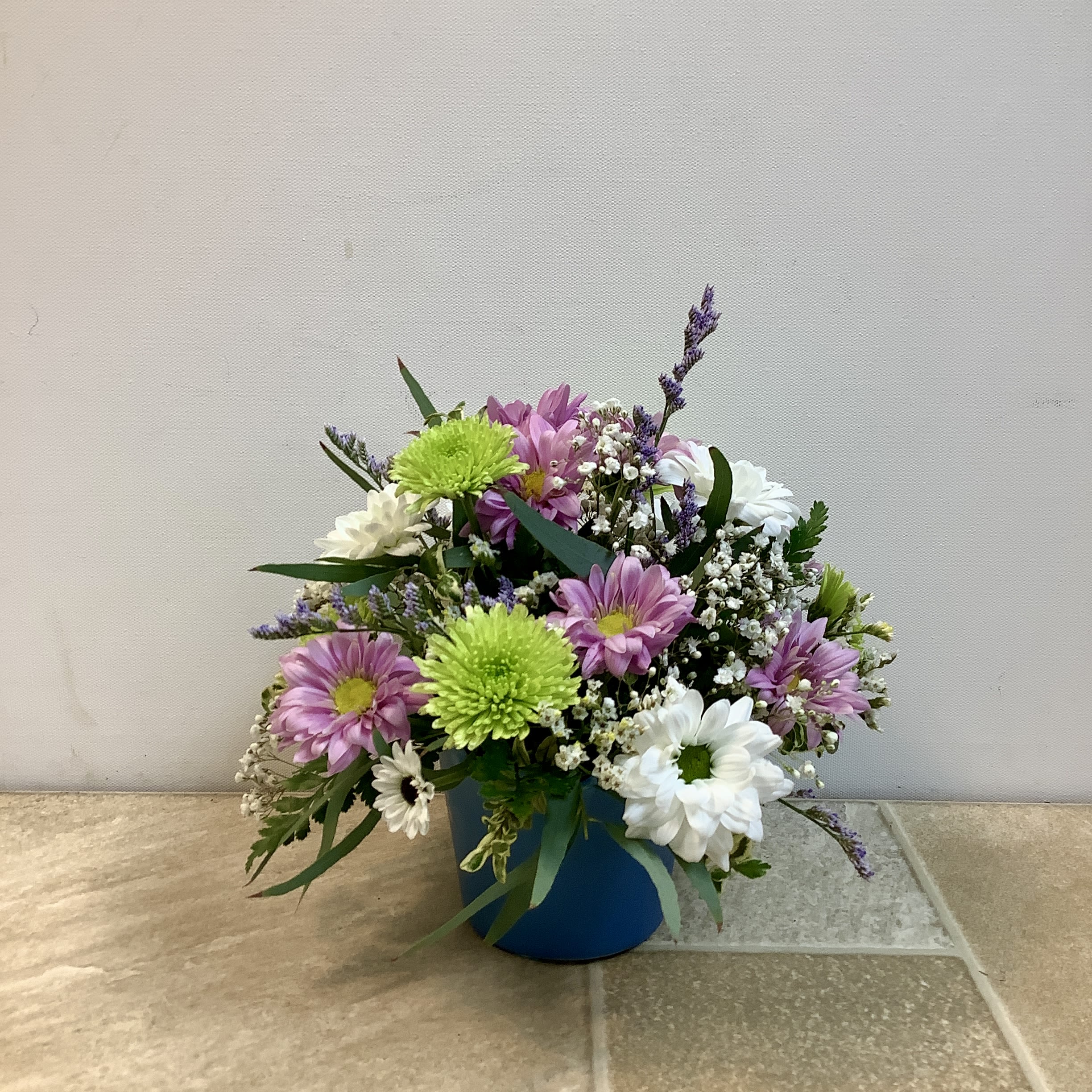 Sweet and Petite - This petite bouquet is a sweet and cheerful floral arrangement displayed in a simple blue container. It features a charming mix of blooms including soft lavender daisy mums, crisp white daisies, and bright green athos.  The flowers are accented with delicate sprigs of baby’s breath and lush fern greenery, creating a fresh and airy look.  Compact in size and beautifully balanced, this bouquet radiates a light, uplifting feel — perfect for brightening up a desk, bedside table, or as a thoughtful “just because” gift. It measures 10” wide x 8” high.   