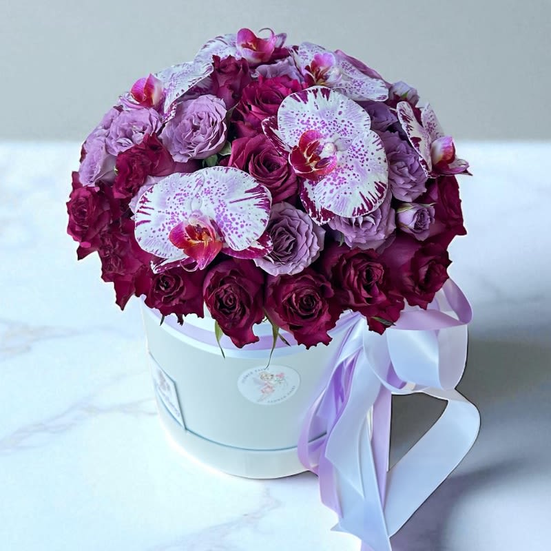 Mystic Amour Flower Box #81 Mystic Amour Flower Box #81 - Deep, velvety purple roses and soft lavender spray roses mingle with exotic cymbidium orchids, all arranged in a crisp white hat box, crafted to make a lasting impression. A luxurious gift that exudes romance and mystery—perfect for someone who appreciates elegance in every detail. Ideal for marking meaningful moments with style. Please note: the exact blooms may vary depending on availability, but the overall color palette and luxurious feel will remain the same.